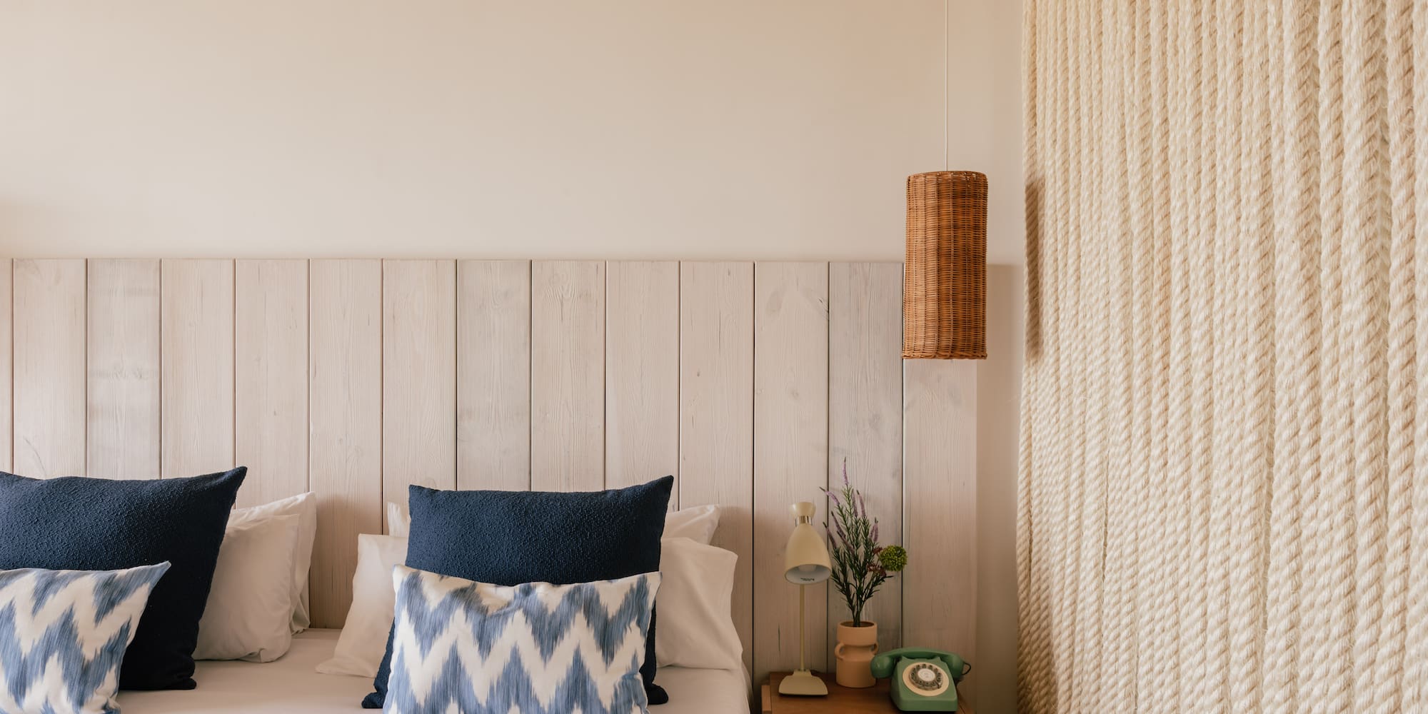 Habitación estilo costero con cama, cabecero madera, lámpara mimbre y pared de cuerda.