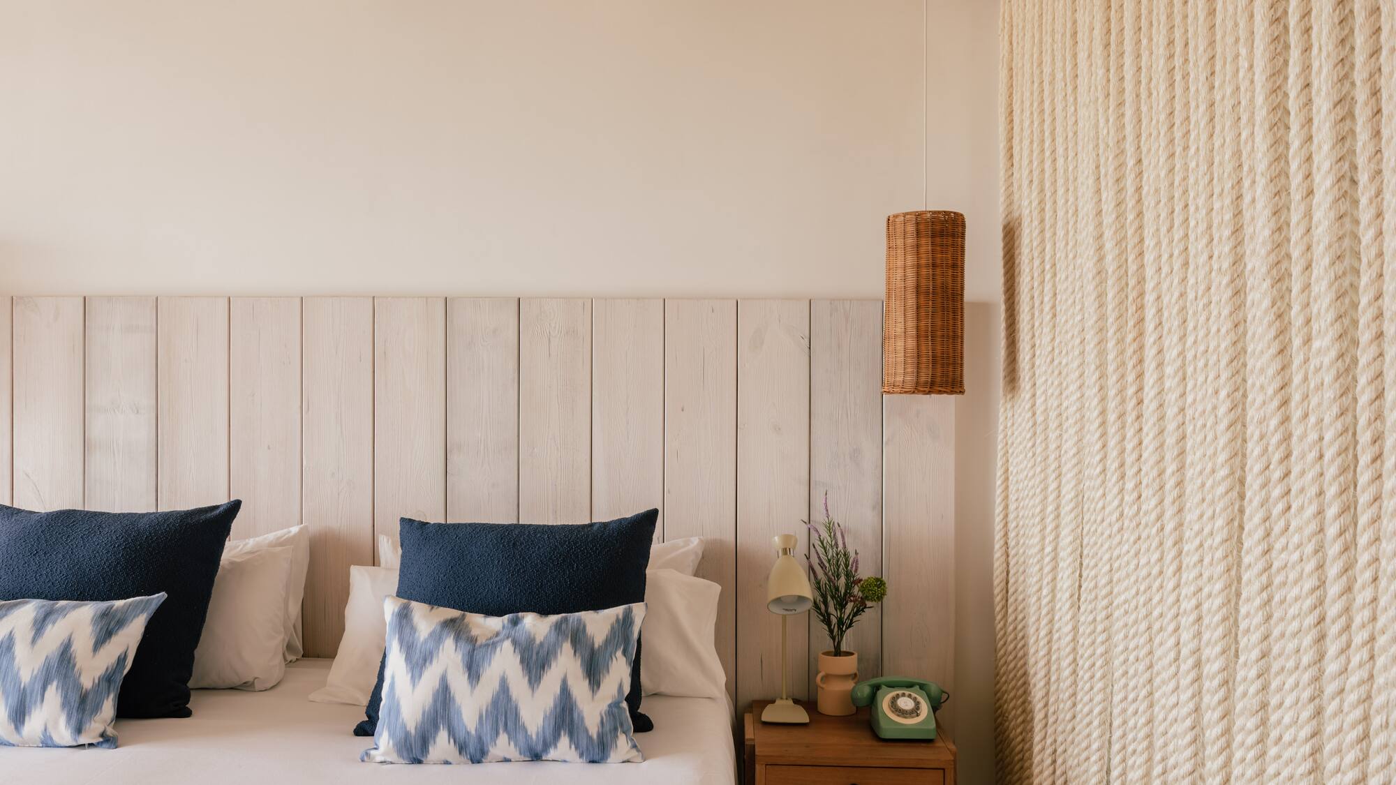 Habitación estilo costero con cama, cabecero madera, lámpara mimbre y pared de cuerda.