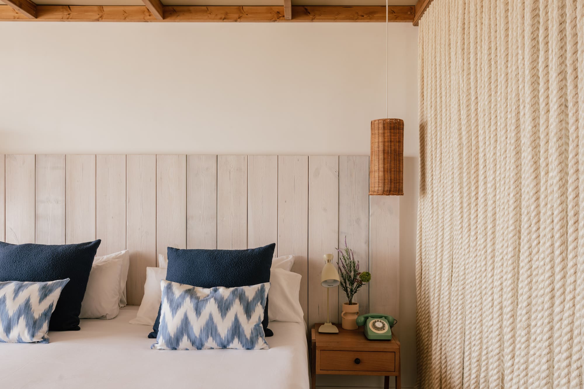 Habitación estilo costero con cama, cabecero madera, lámpara mimbre y pared de cuerda.