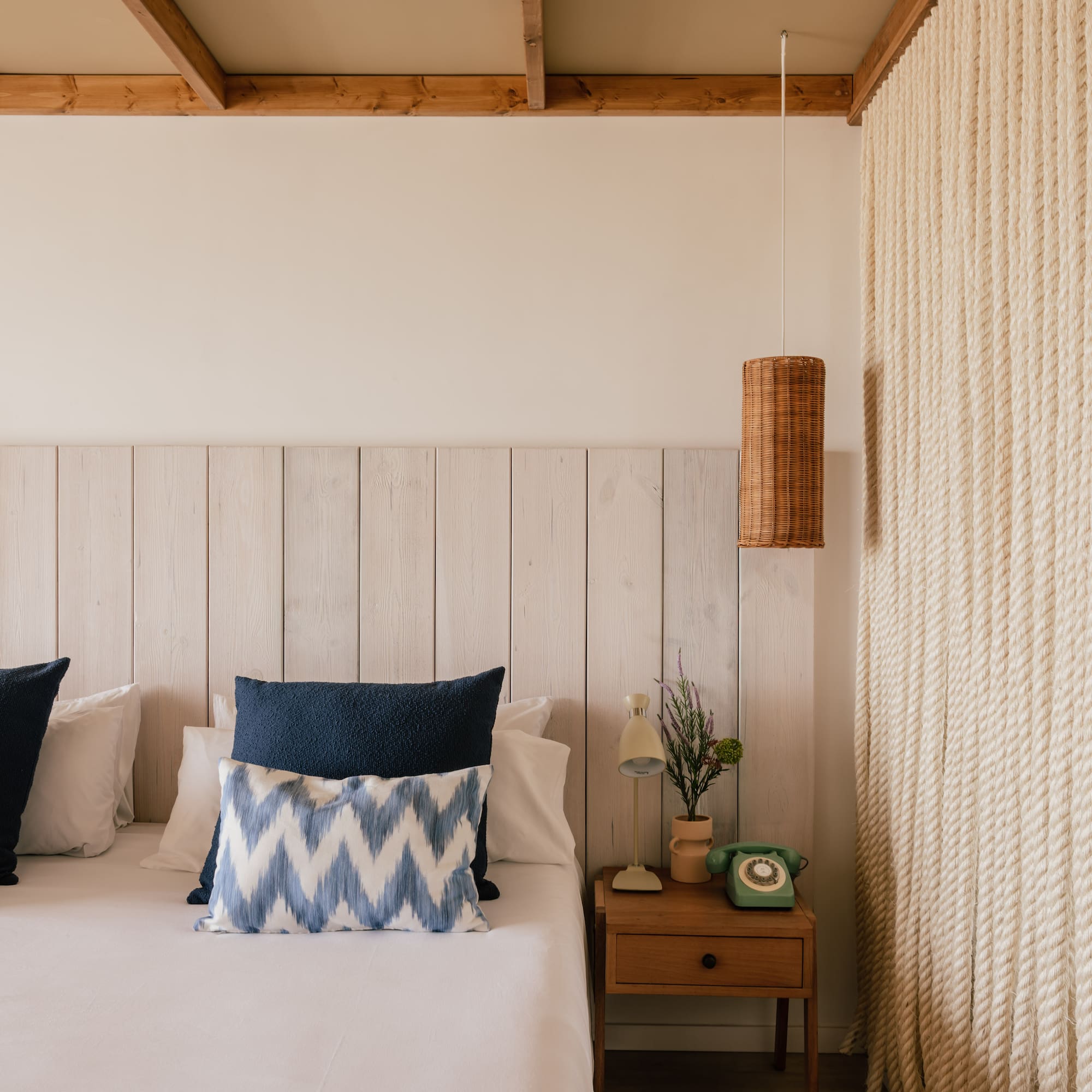 Habitación estilo costero con cama, cabecero madera, lámpara mimbre y pared de cuerda.