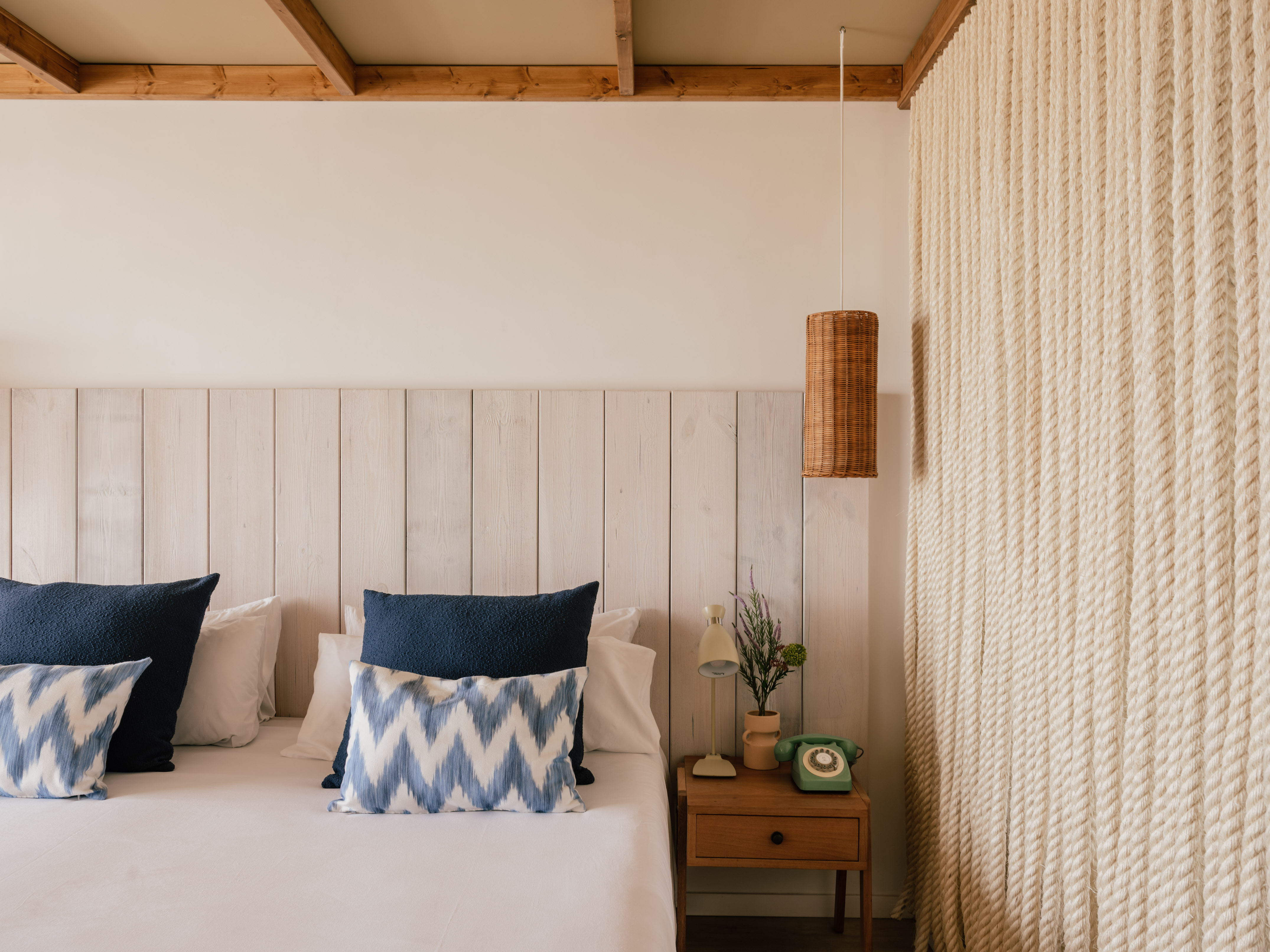 Habitación estilo costero con cama, cabecero madera, lámpara mimbre y pared de cuerda.