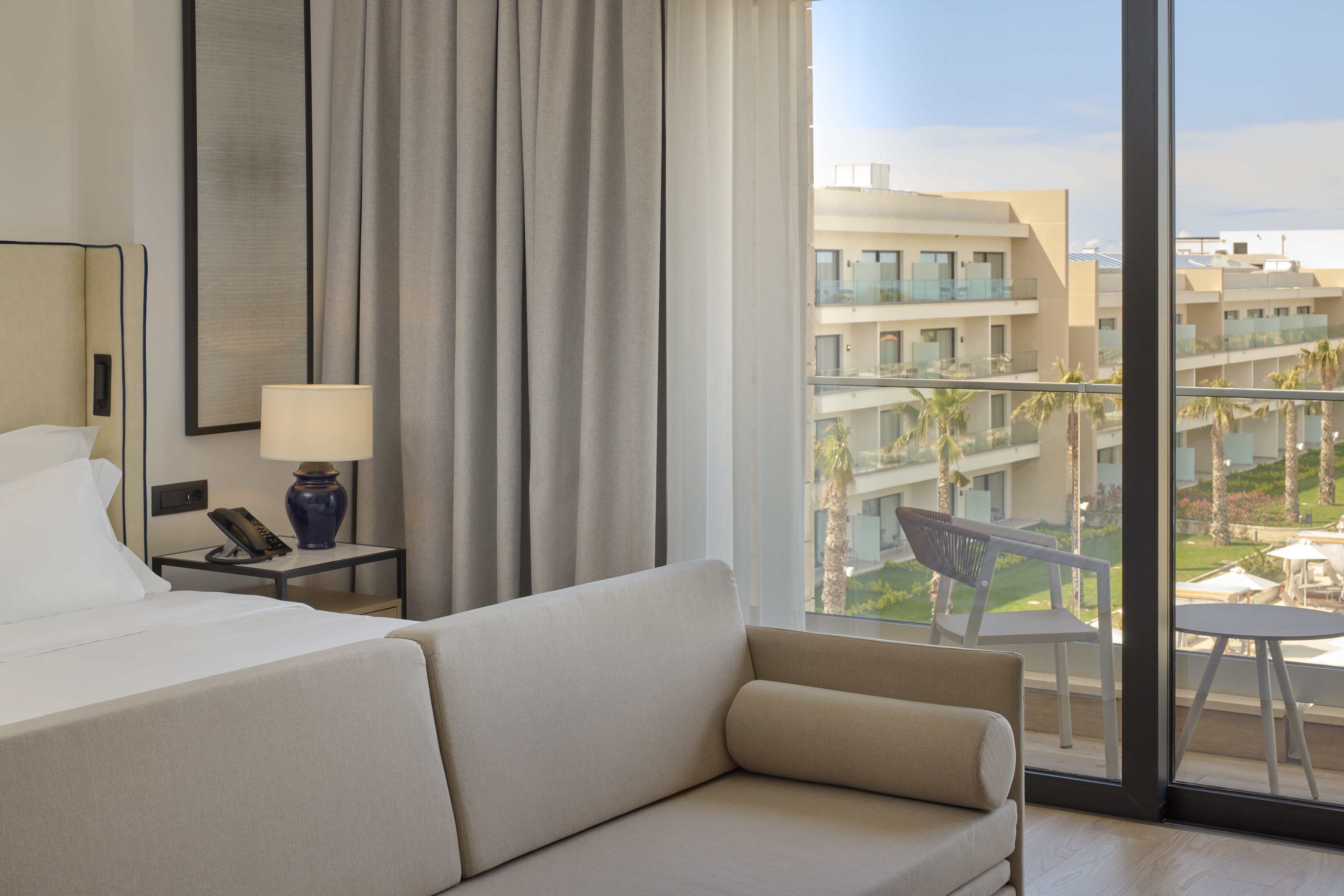 Modern hotel room with sofa, bed, and balcony view of a resort complex.