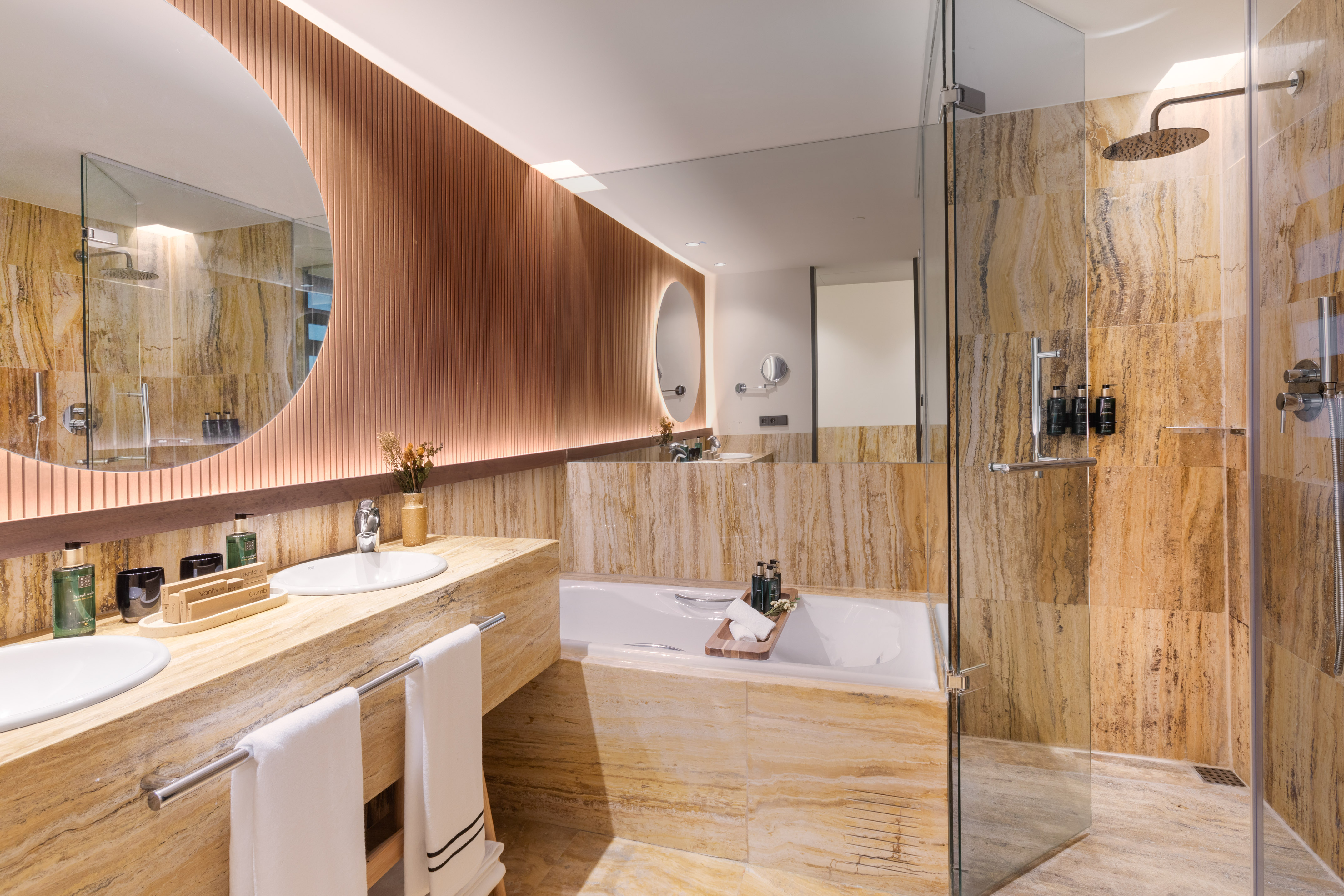Modern bathroom with stone double vanity, circular mirror, bathtub, and glass shower.