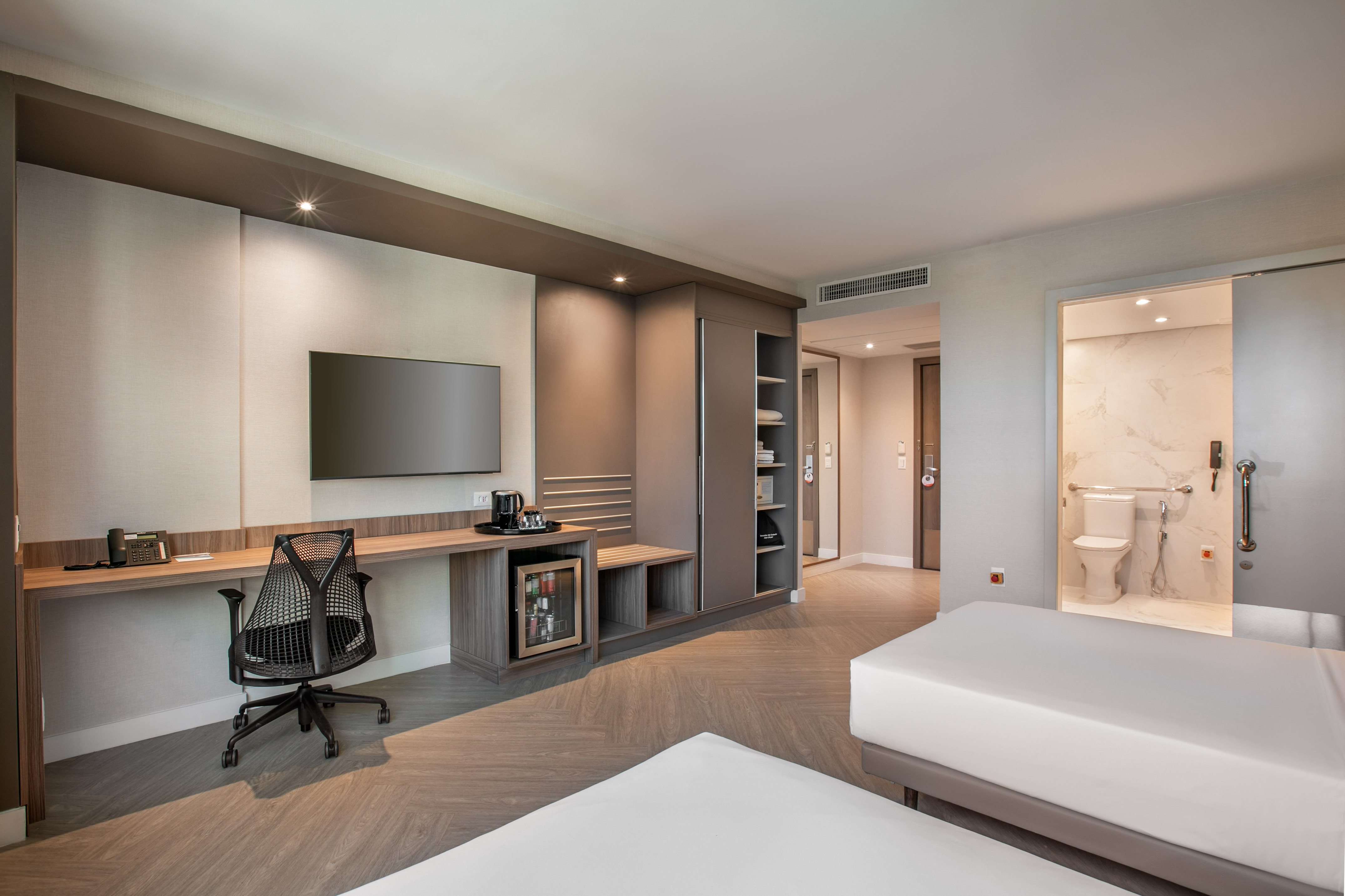 Modern hotel room featuring beds, desk, TV, and accessible bathroom.