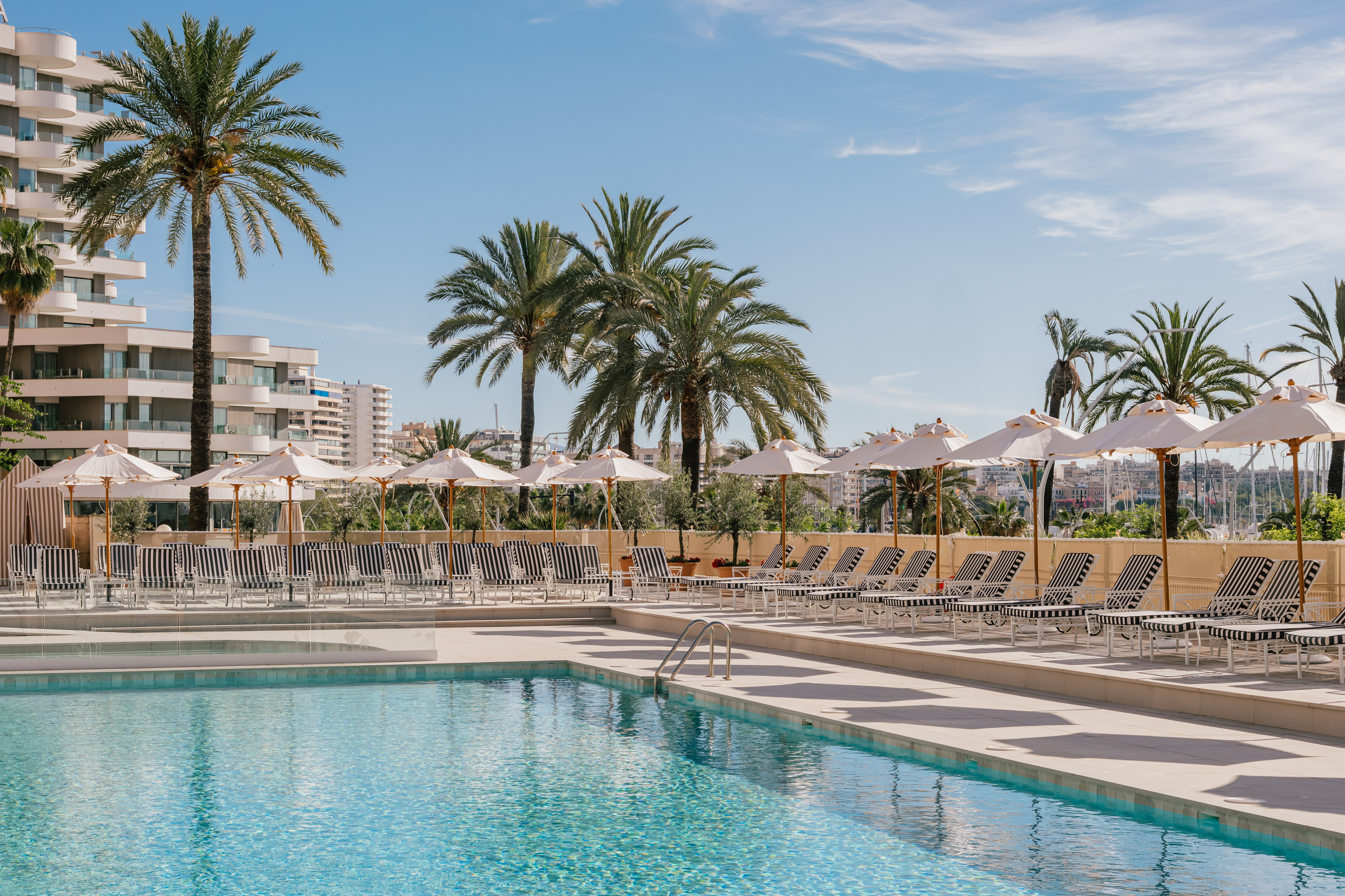 Luxury resort pool with lounge chairs, umbrellas, palm trees, and marina view.