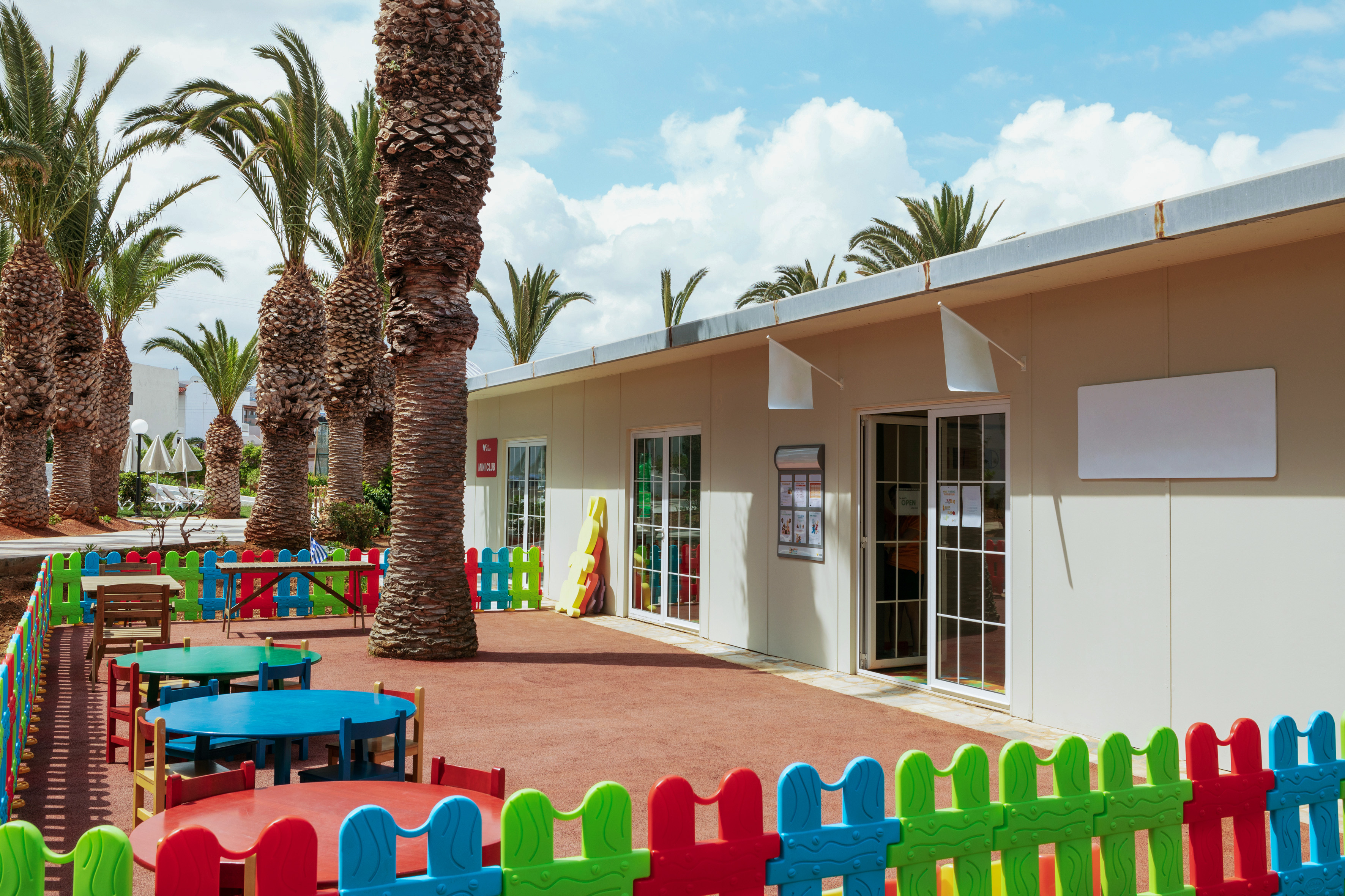 Outdoor mini club play area with colorful fence, tables, chairs, and palm trees.