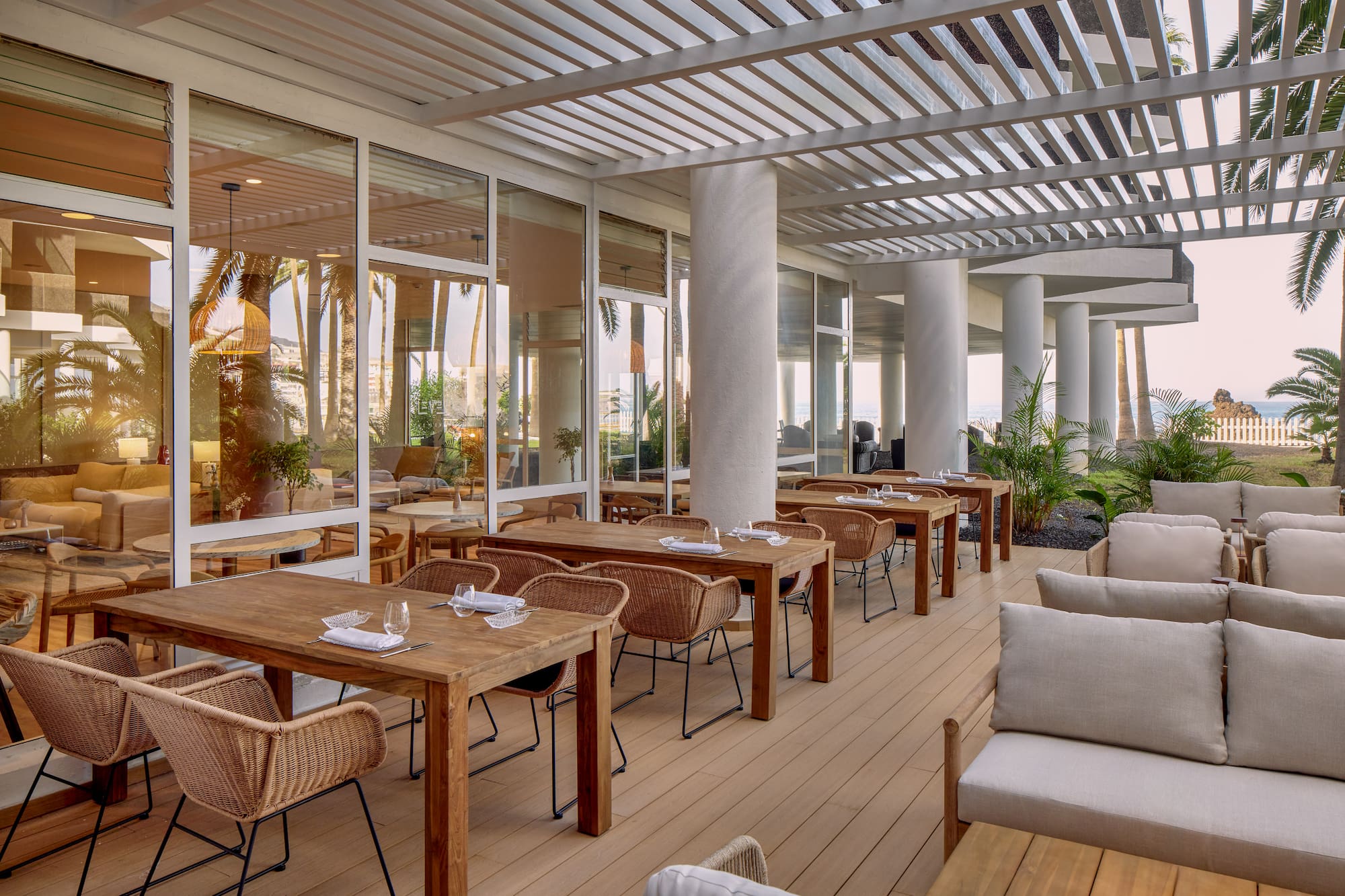 Resort outdoor dining terrace with wooden tables, wicker chairs, ocean view.