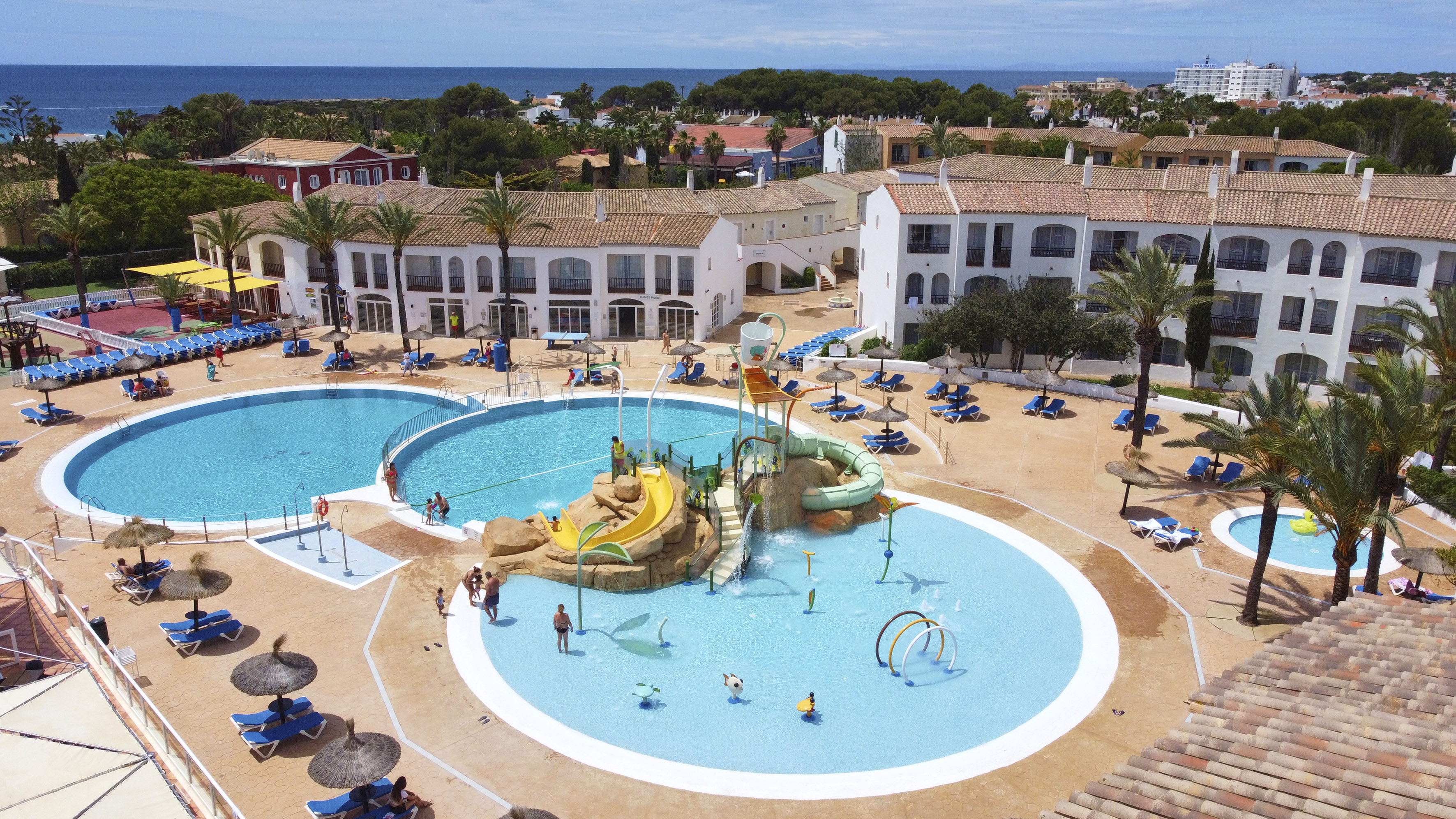 Mediterranean resort pools, water park, palm trees, and distant ocean view.