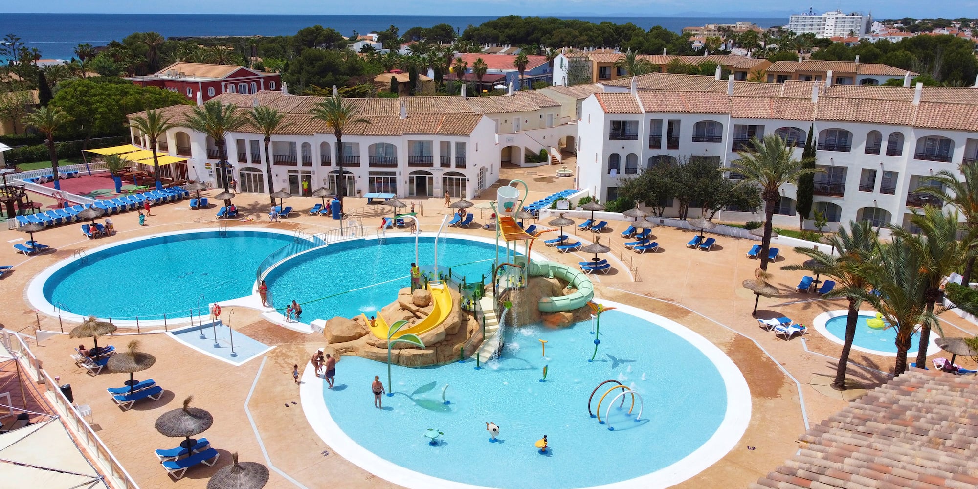 Mediterranean resort pools, water park, palm trees, and distant ocean view.