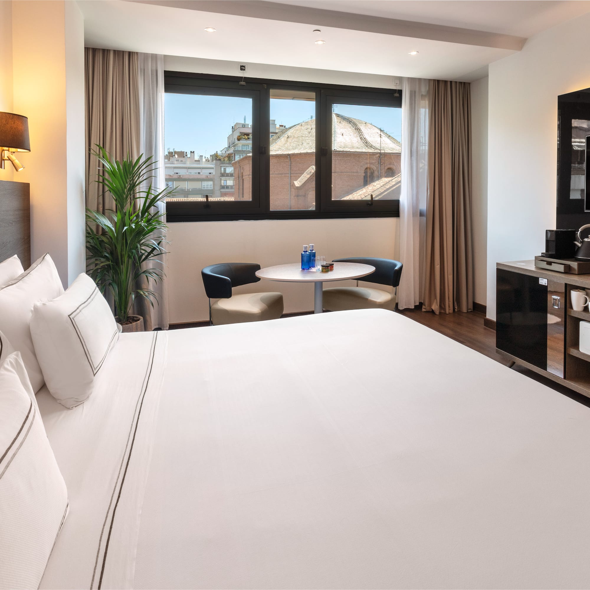 Modern hotel room with comfortable bed, city view window, and minibar.