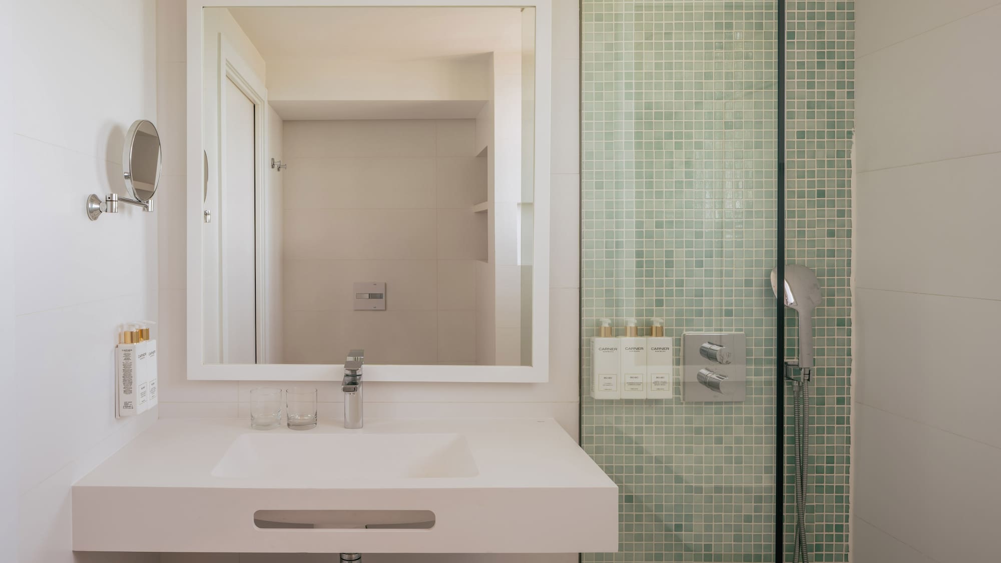 Modern hotel bathroom vanity with mirror and green mosaic tile shower.