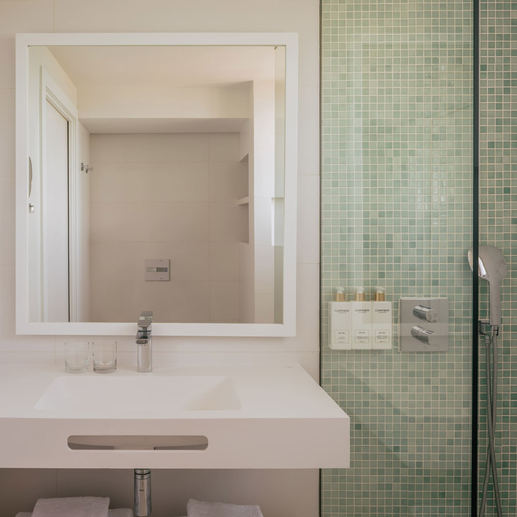 Modern hotel bathroom vanity with mirror and green mosaic tile shower.