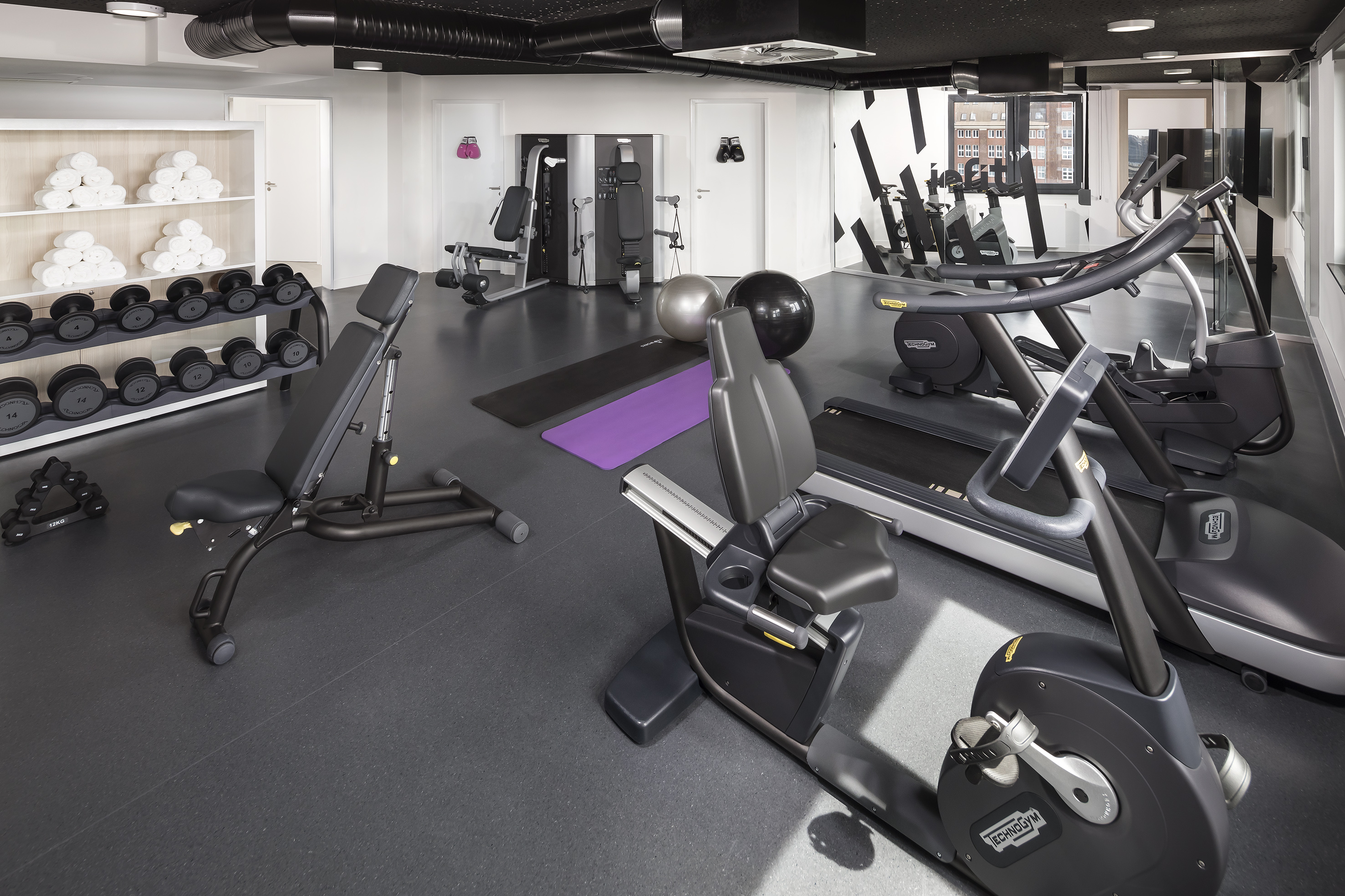 Modern hotel gym with dumbbells, treadmill, exercise bike, yoga mats, and white towels.