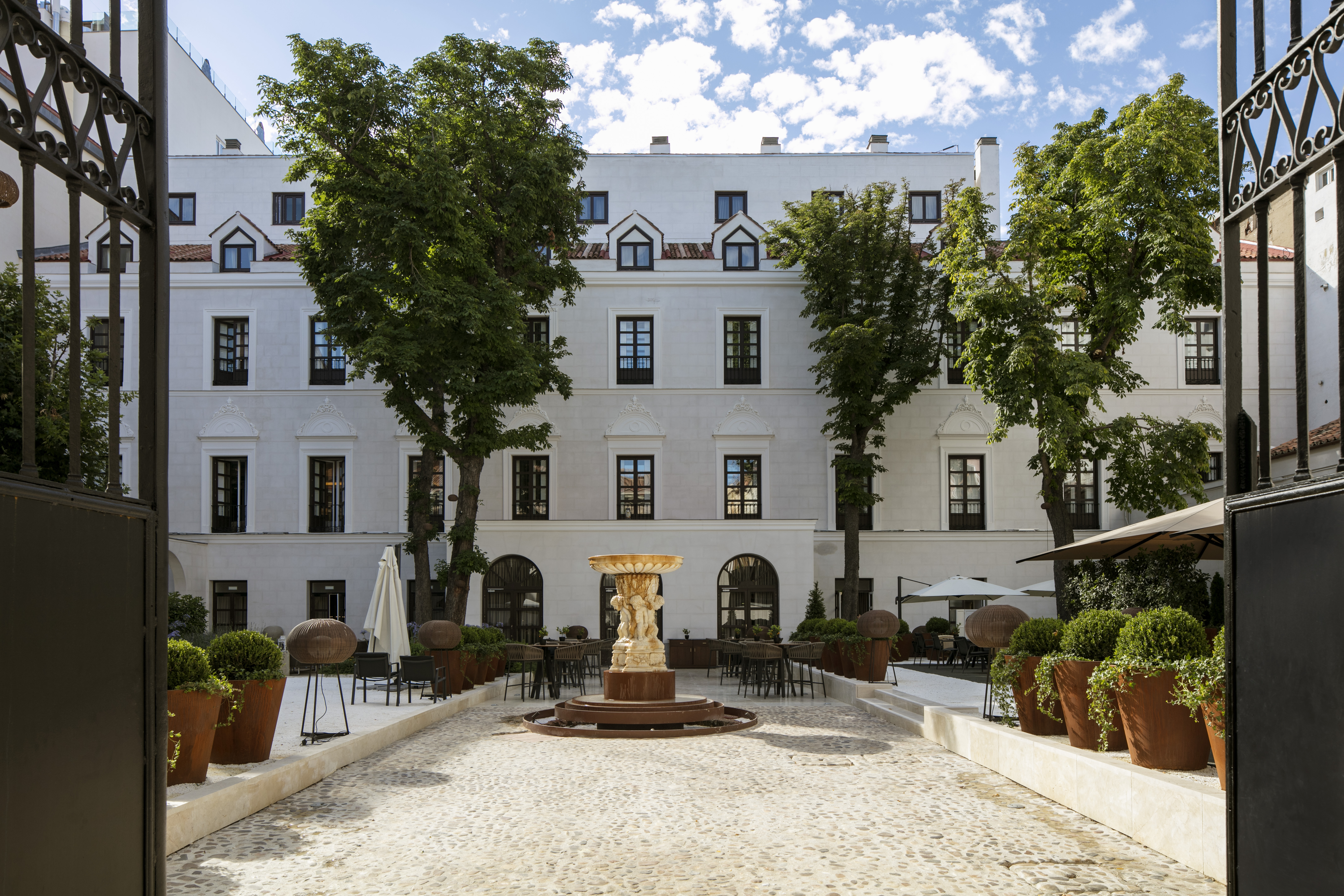 Hotel courtyard with stone fountain, outdoor seating, green trees, white building.