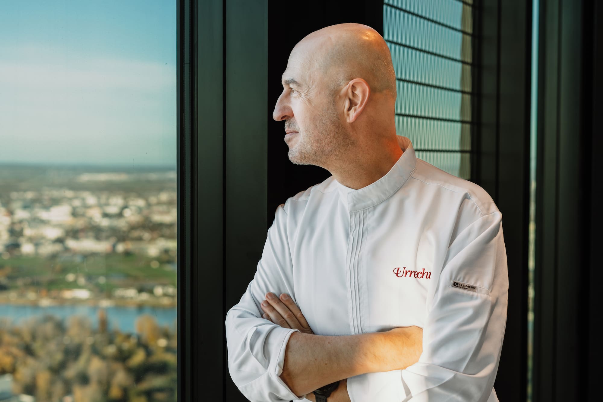 Chef Urrechu in white jacket looks out window at city skyline.