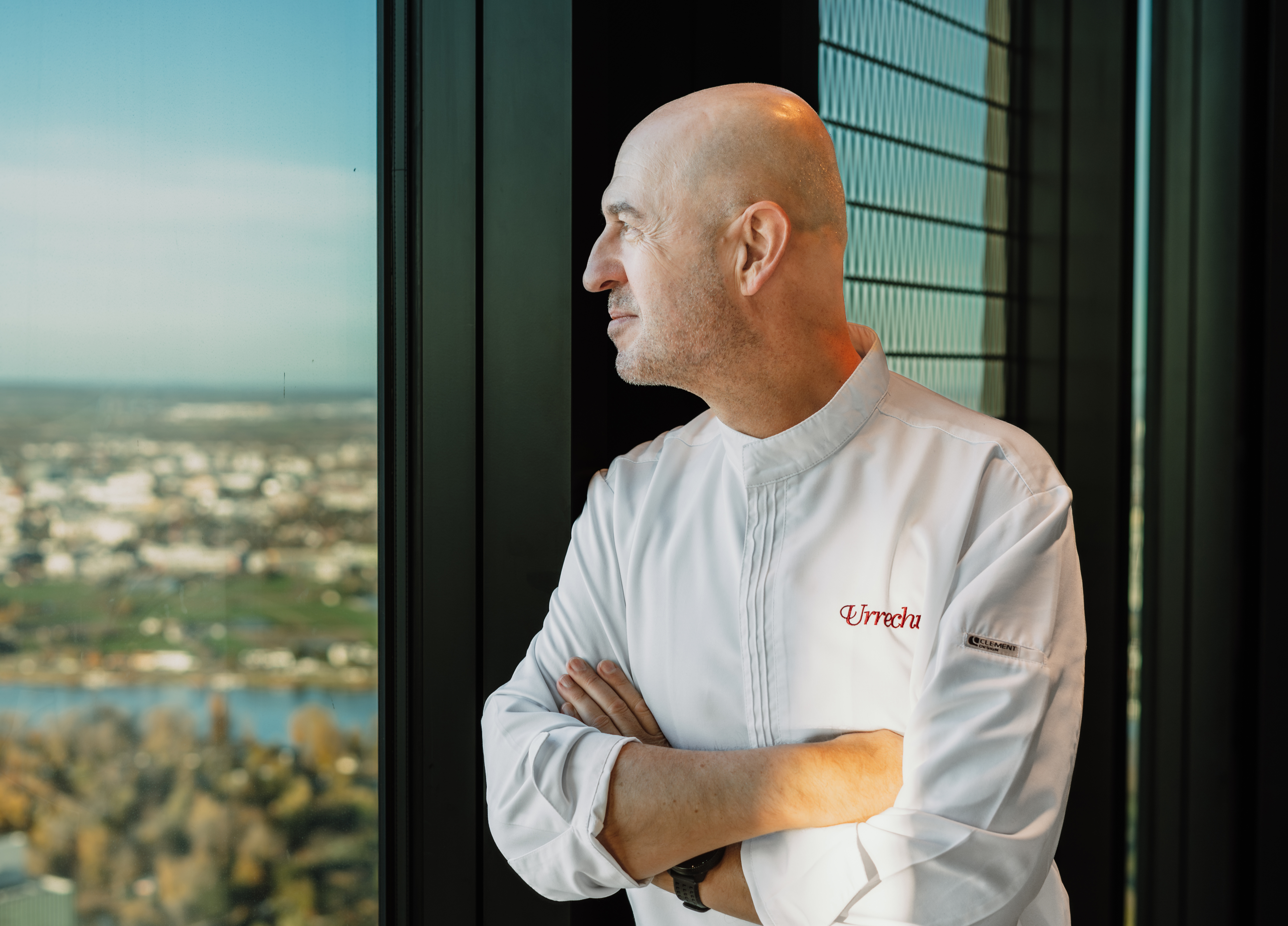 Chef Urrechu in white jacket looks out window at city skyline.