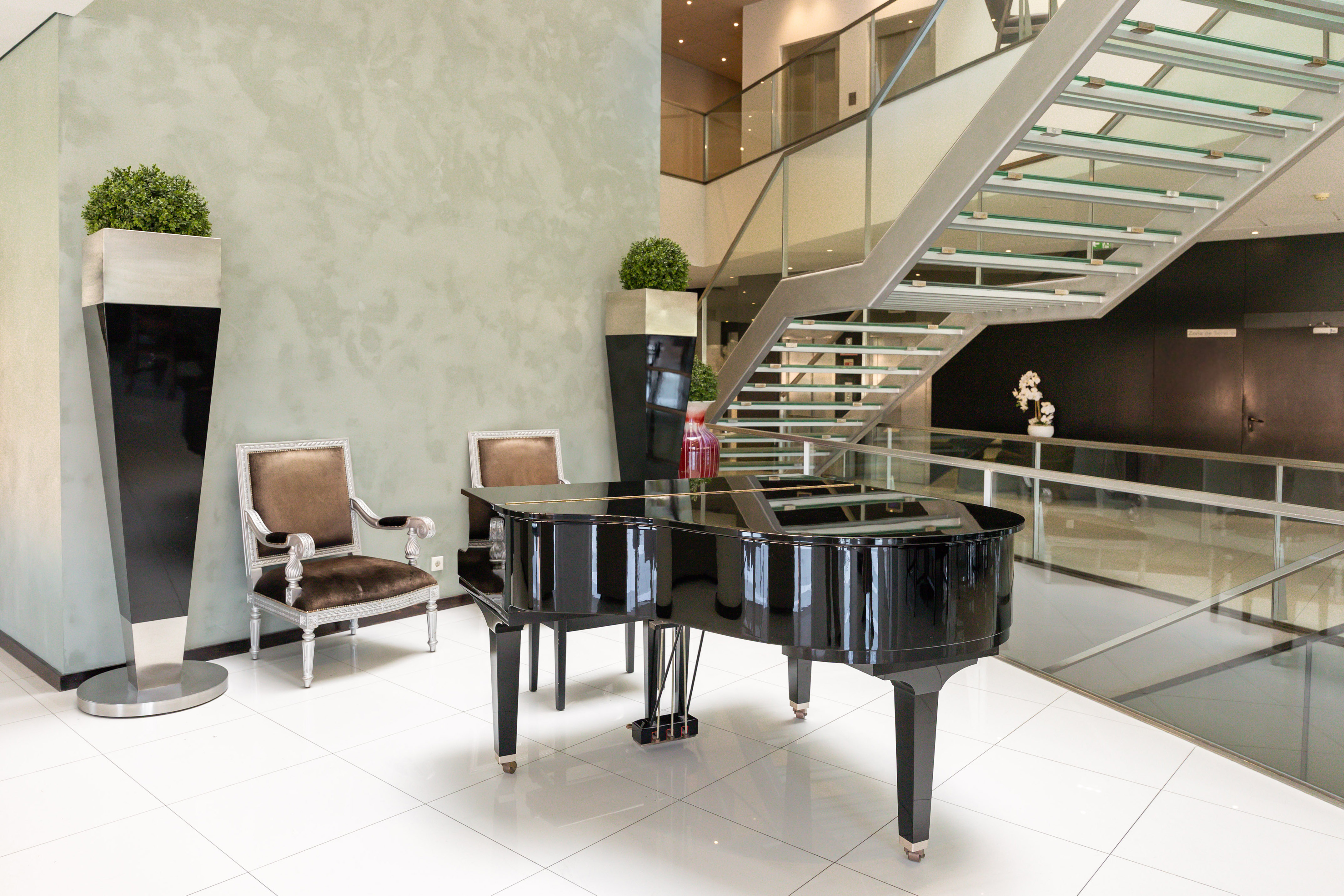 Modern hotel lobby with grand piano, glass staircase, and velvet chairs.
