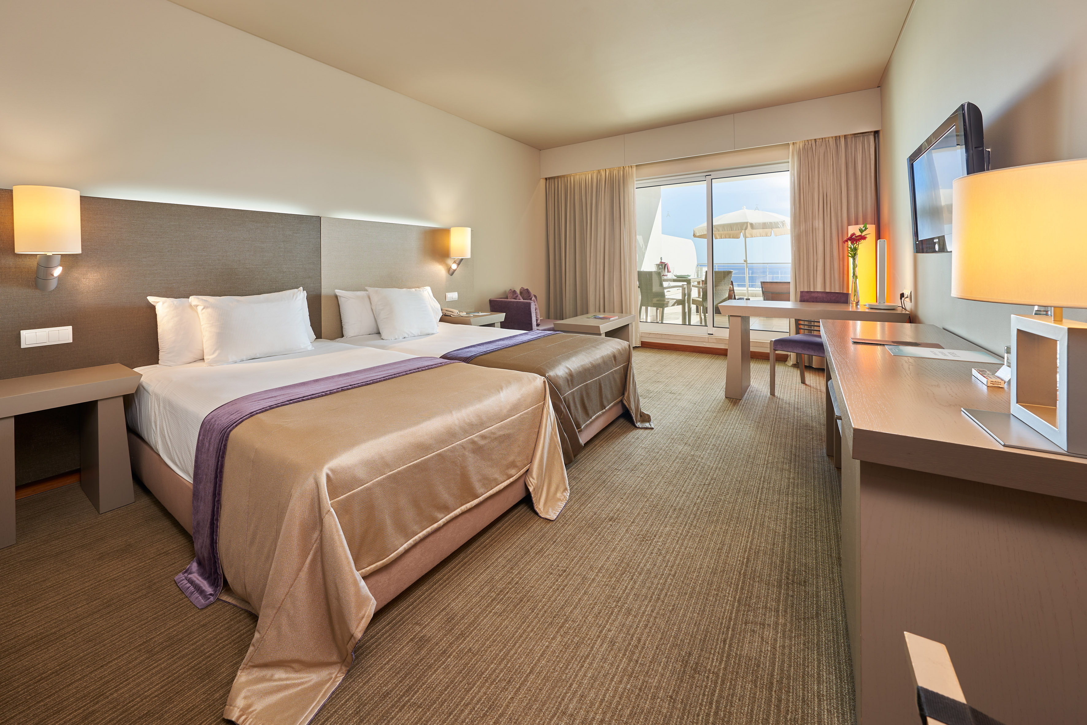 Spacious hotel room with two beds, desk, TV, and balcony with ocean view.