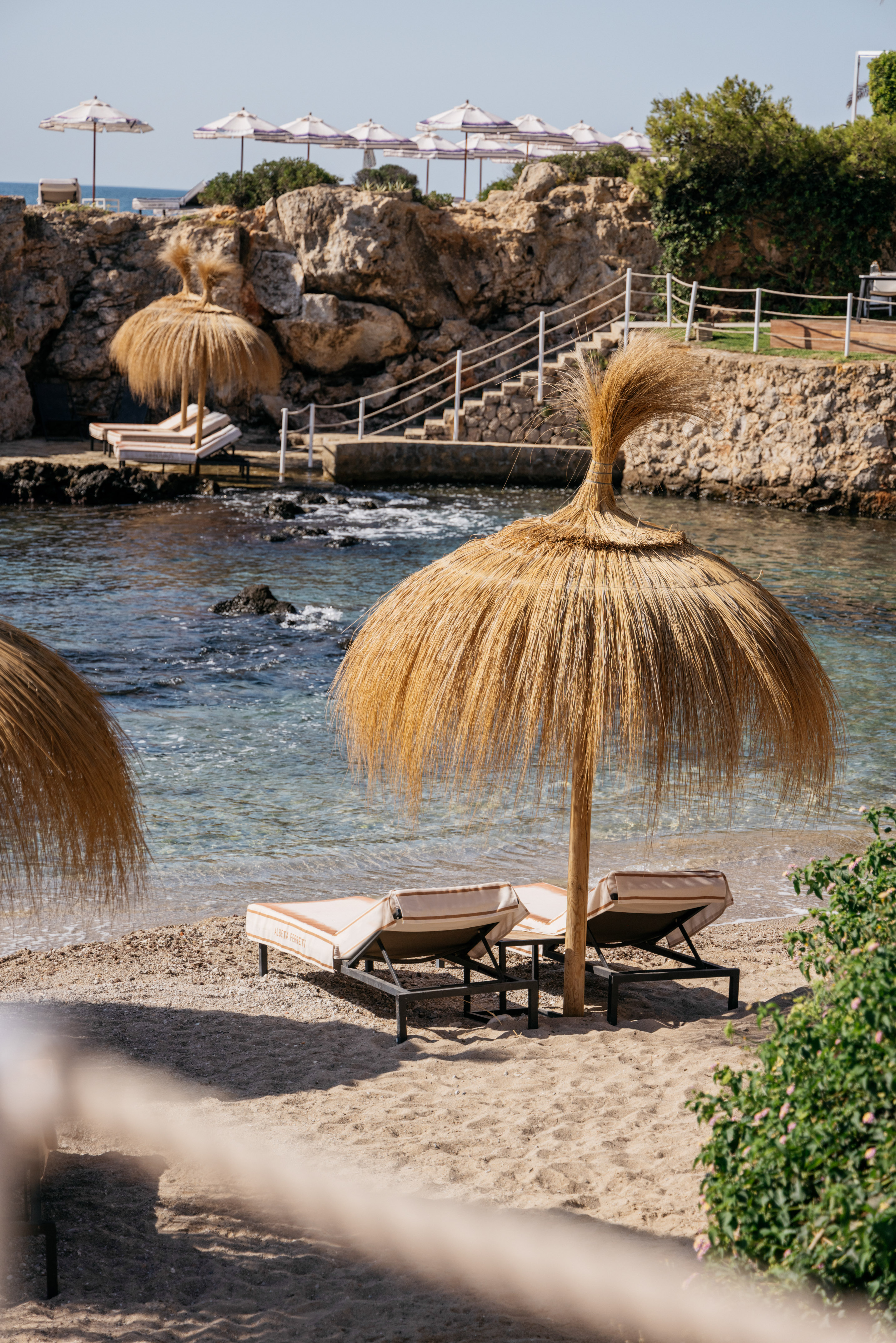 Luxury beach resort with straw umbrellas, sunbeds, clear sea, rocky cliffs.