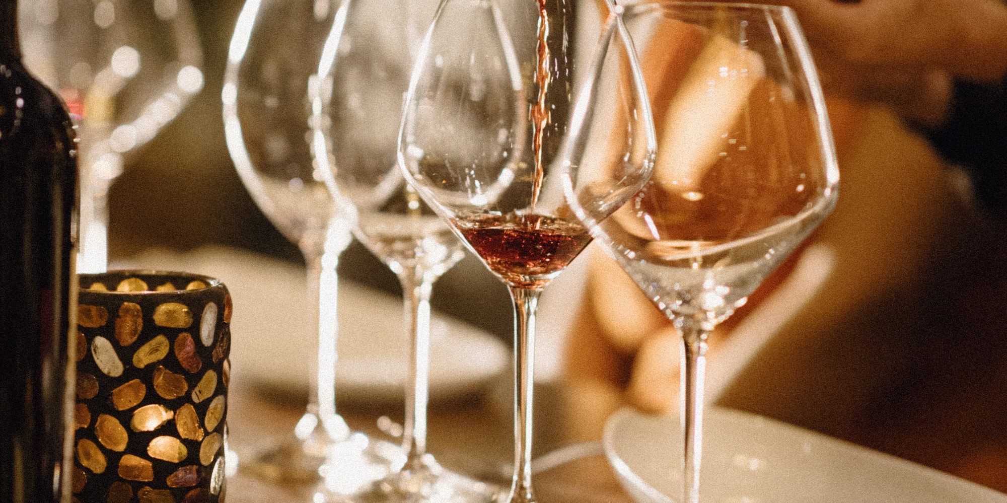 Red wine pouring into a glass, with cheese board and candlelit table setting.