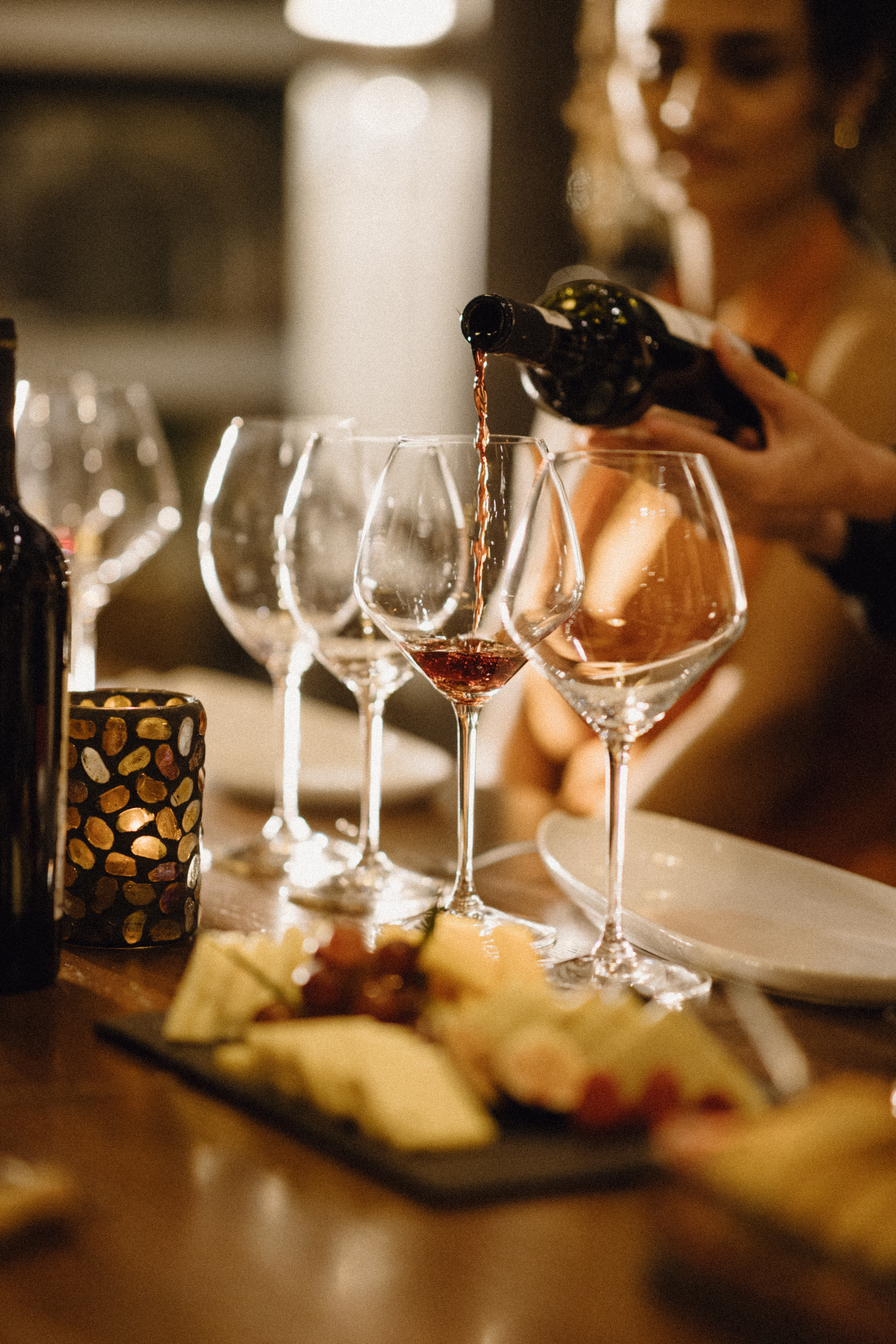Red wine pouring into a glass, with cheese board and candlelit table setting.