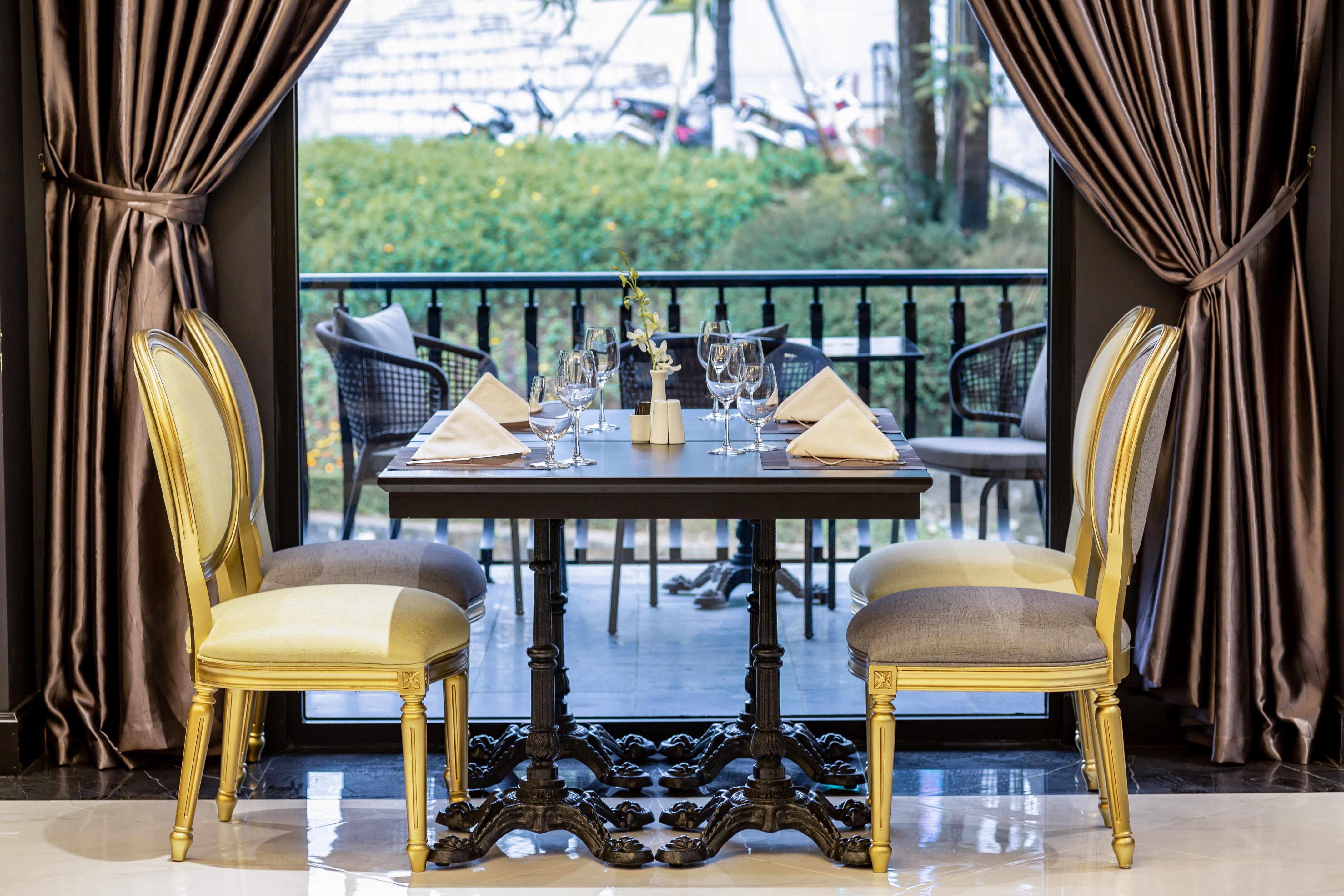 Elegant restaurant dining table, golden chairs, wine glasses, balcony garden view.