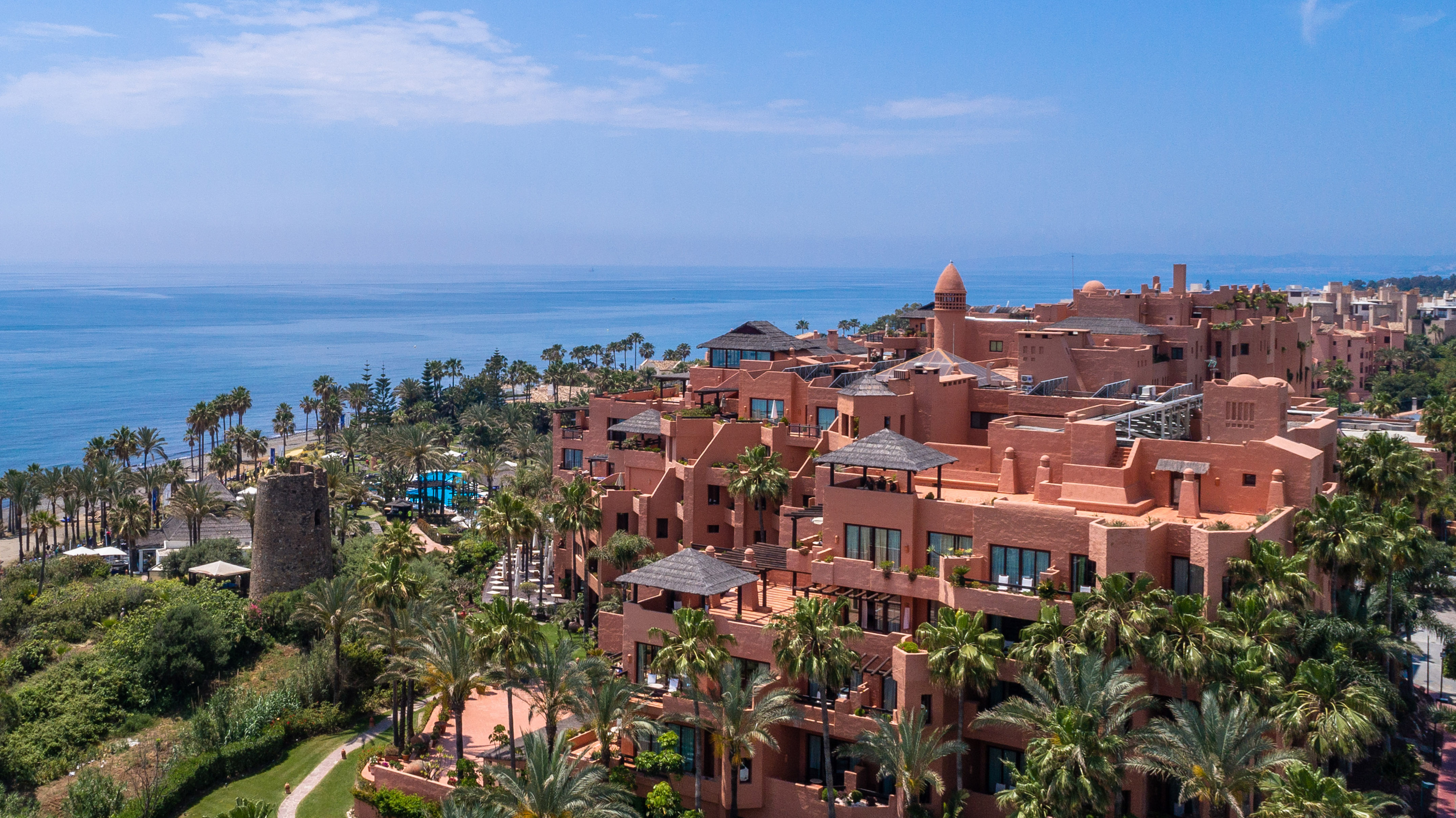 Aerial view of luxury coastal resort with terracotta buildings and palm trees.