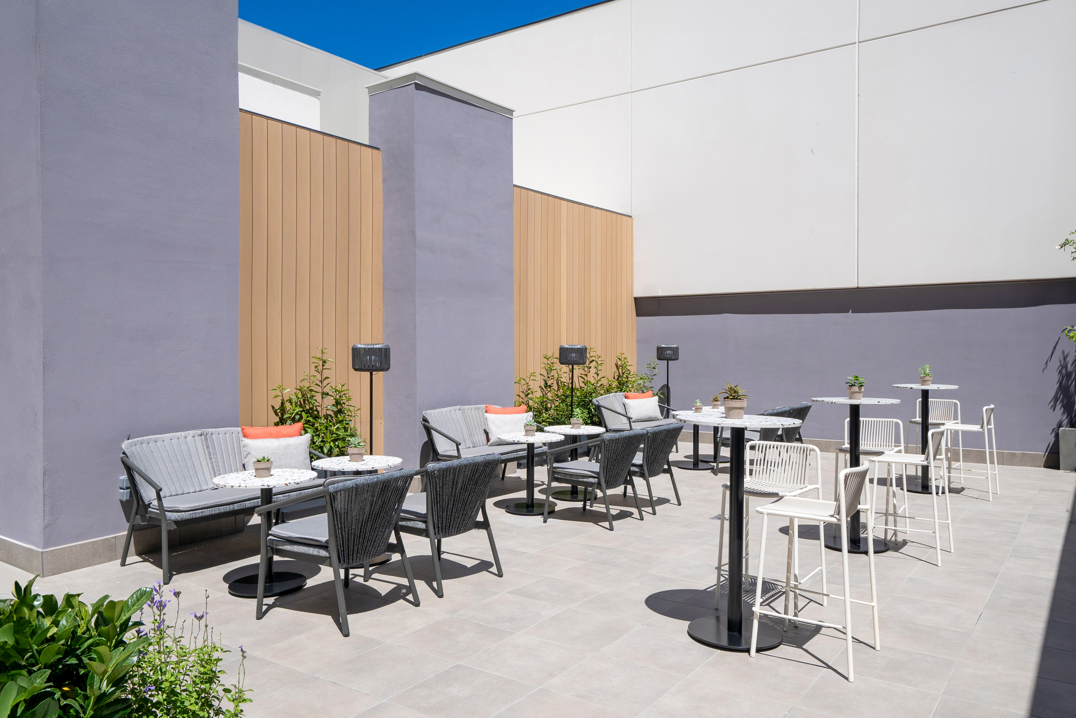 Modern outdoor patio lounge area with grey seating, bar tables, and greenery.