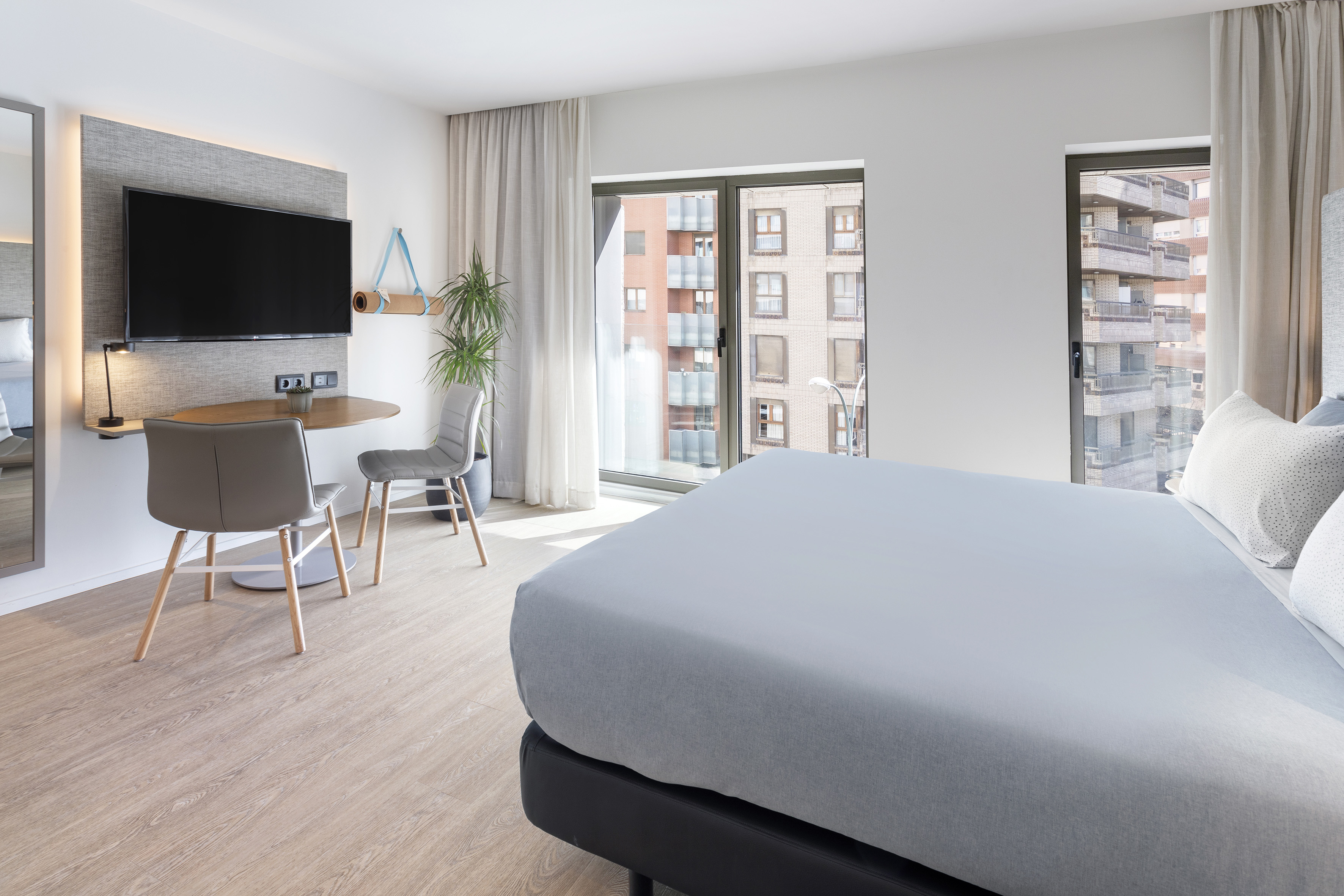 Modern hotel bedroom with large bed, city view windows, mounted TV, and work desk.