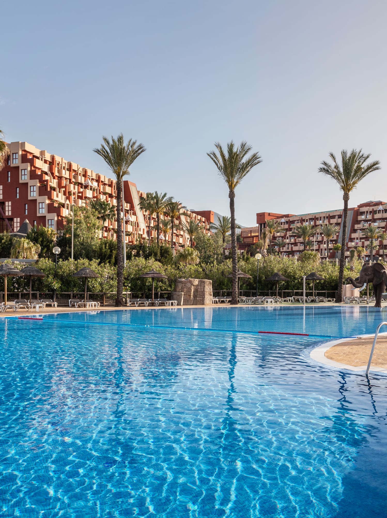 Tropical resort pool with palm trees, multi-story hotels, and elephant sculptures.