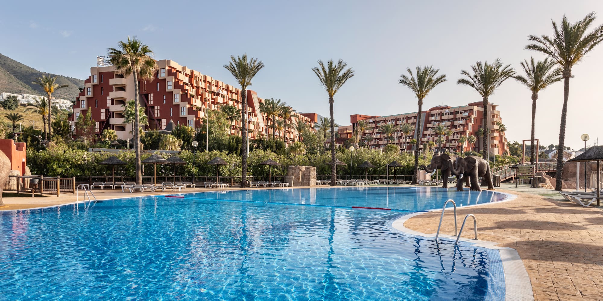 Sunny resort pool, palm trees, elephant statues, and red hotel buildings.