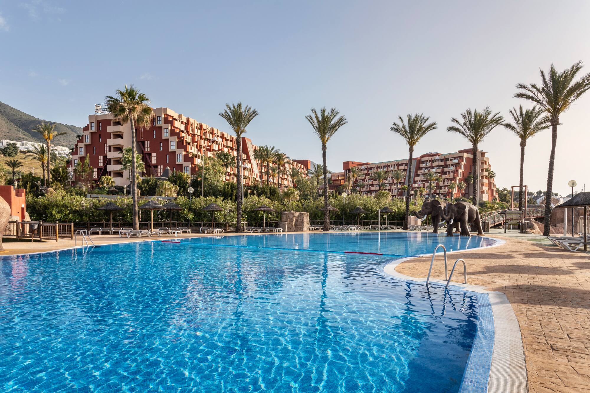 Sunny resort pool, palm trees, elephant statues, and red hotel buildings.