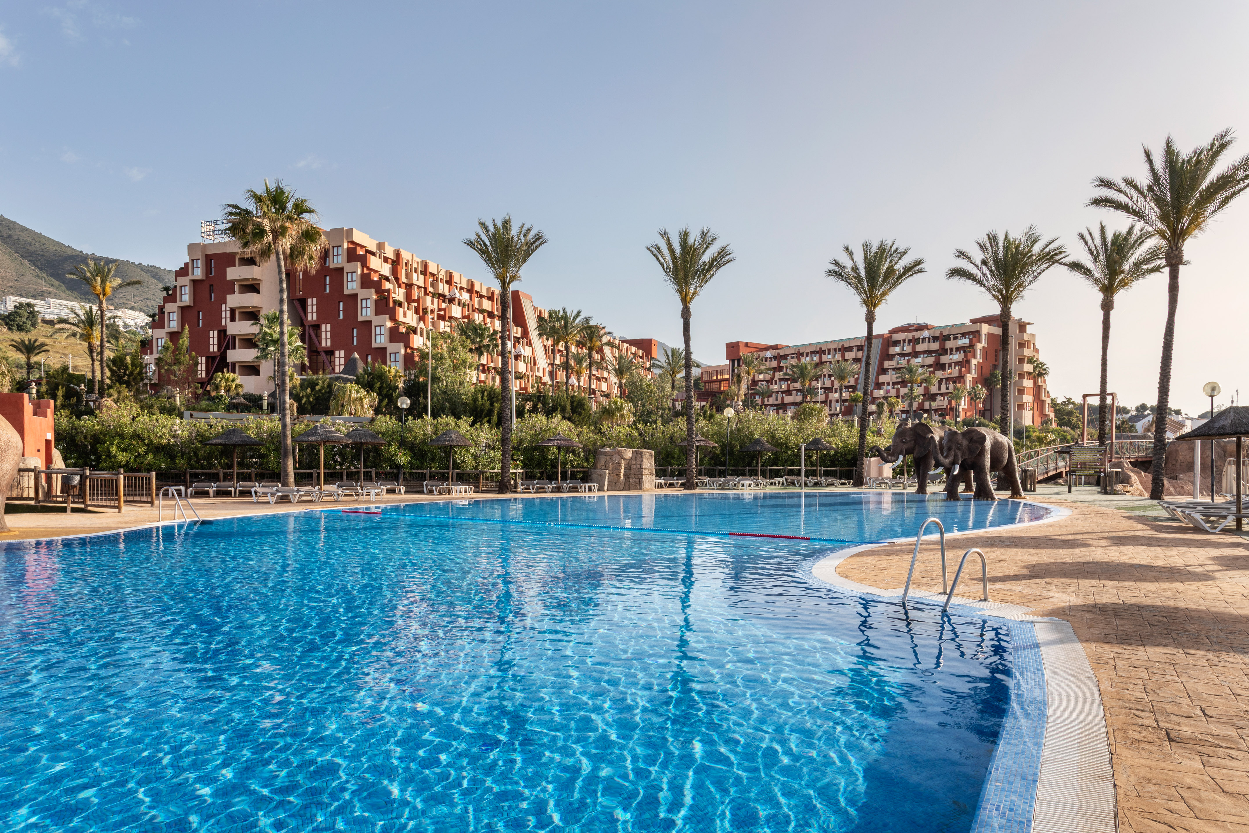 Tropical resort pool with palm trees, multi-story hotels, and elephant sculptures.