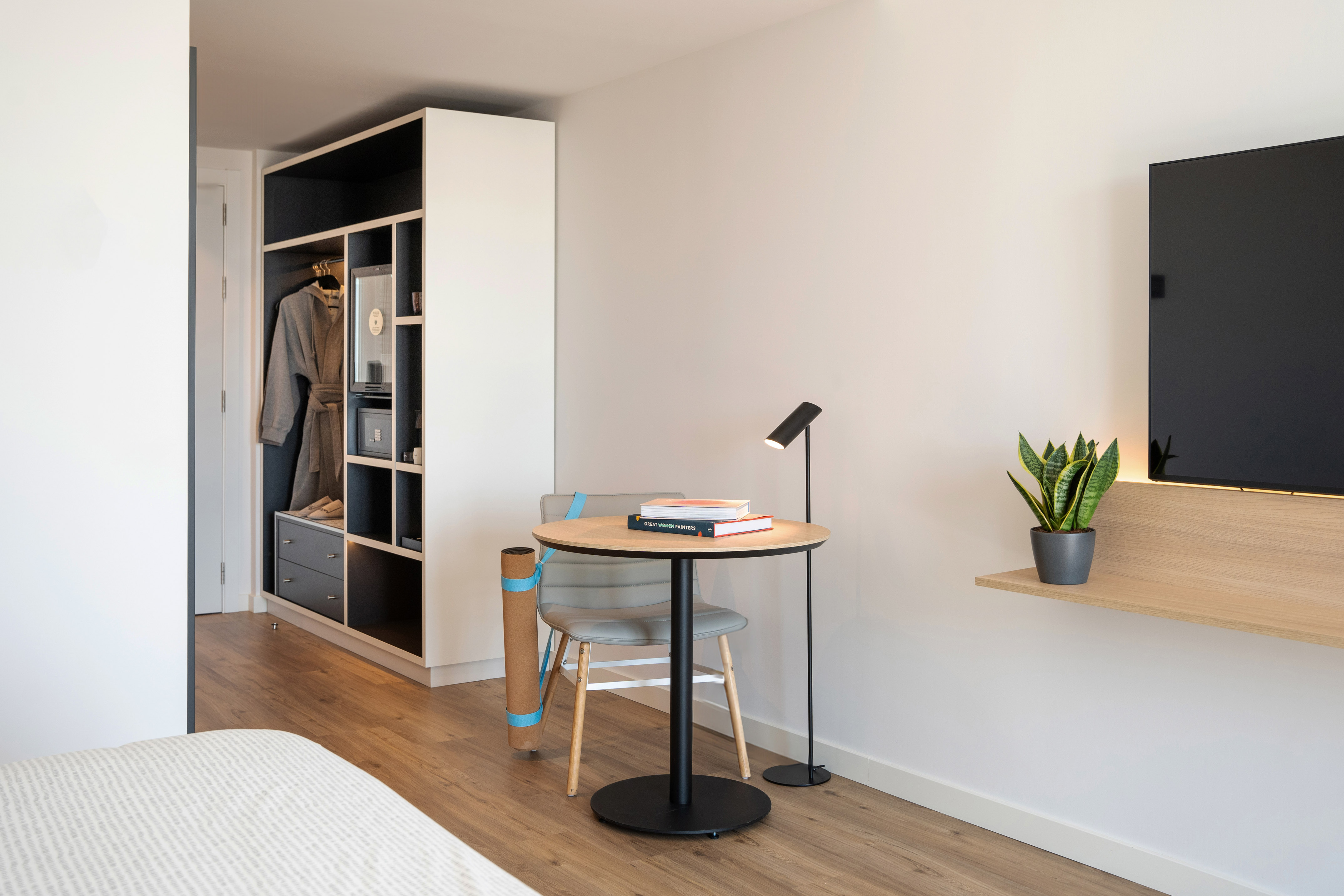 Modern hotel room: open wardrobe, desk area, wall TV, and plant.