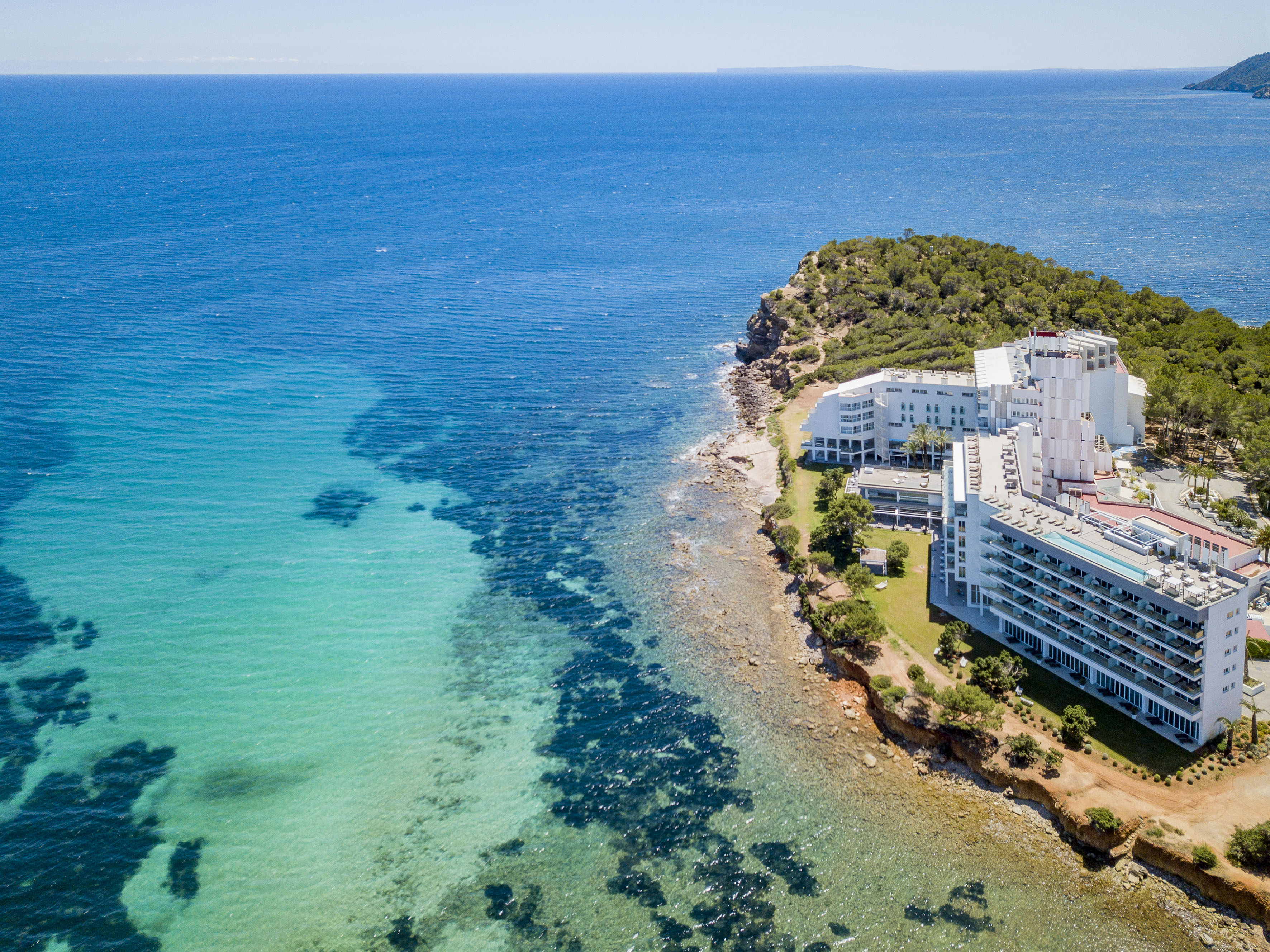 Aerial view: White resort hotel on rocky coastline with clear turquoise ocean.