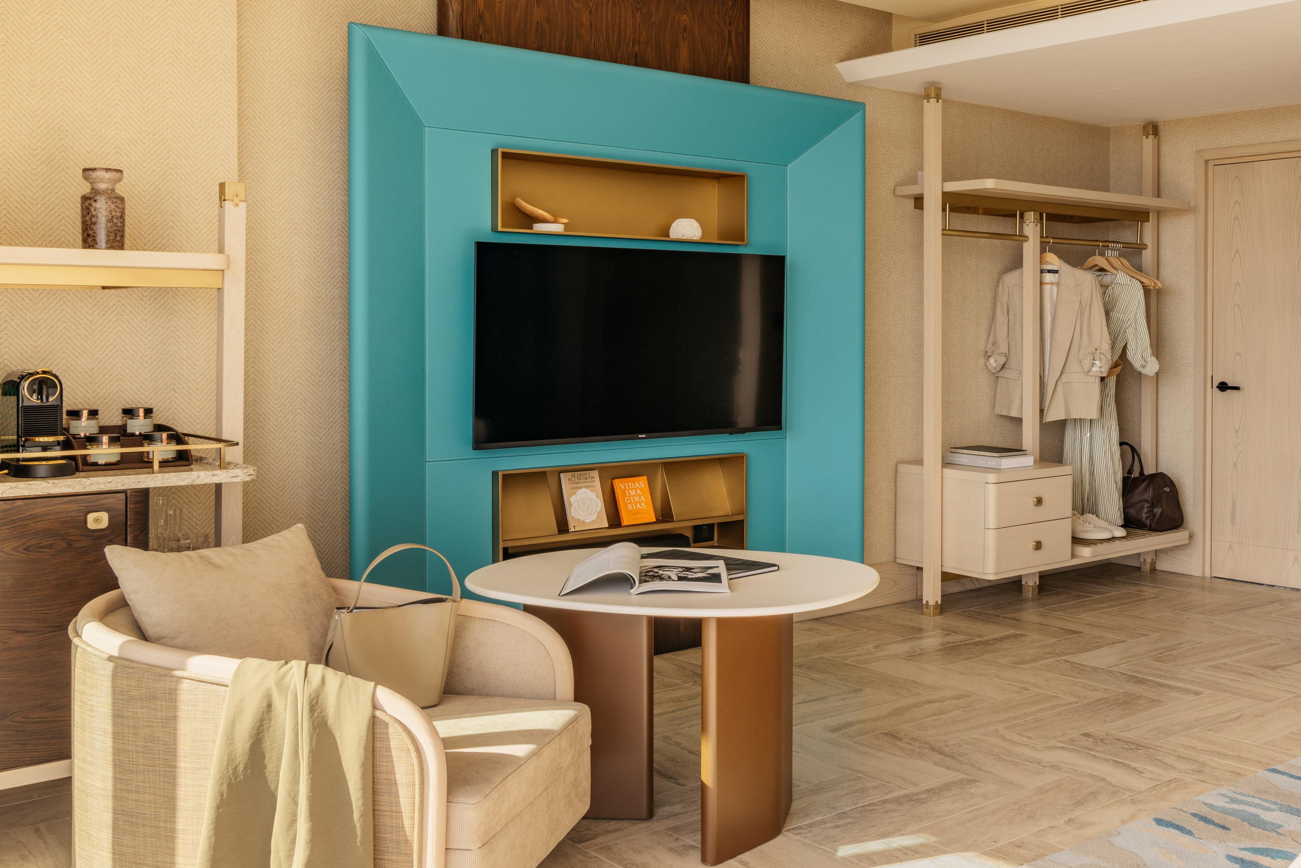 Stylish hotel room with teal media wall, coffee bar, armchair, and open closet.