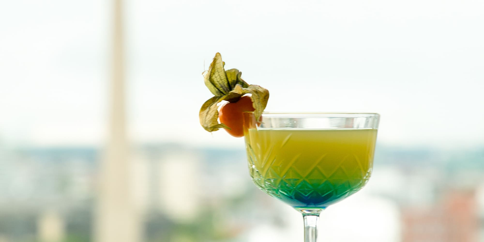 Gradient cocktail with physalis garnish, on table with blurred city tower view.