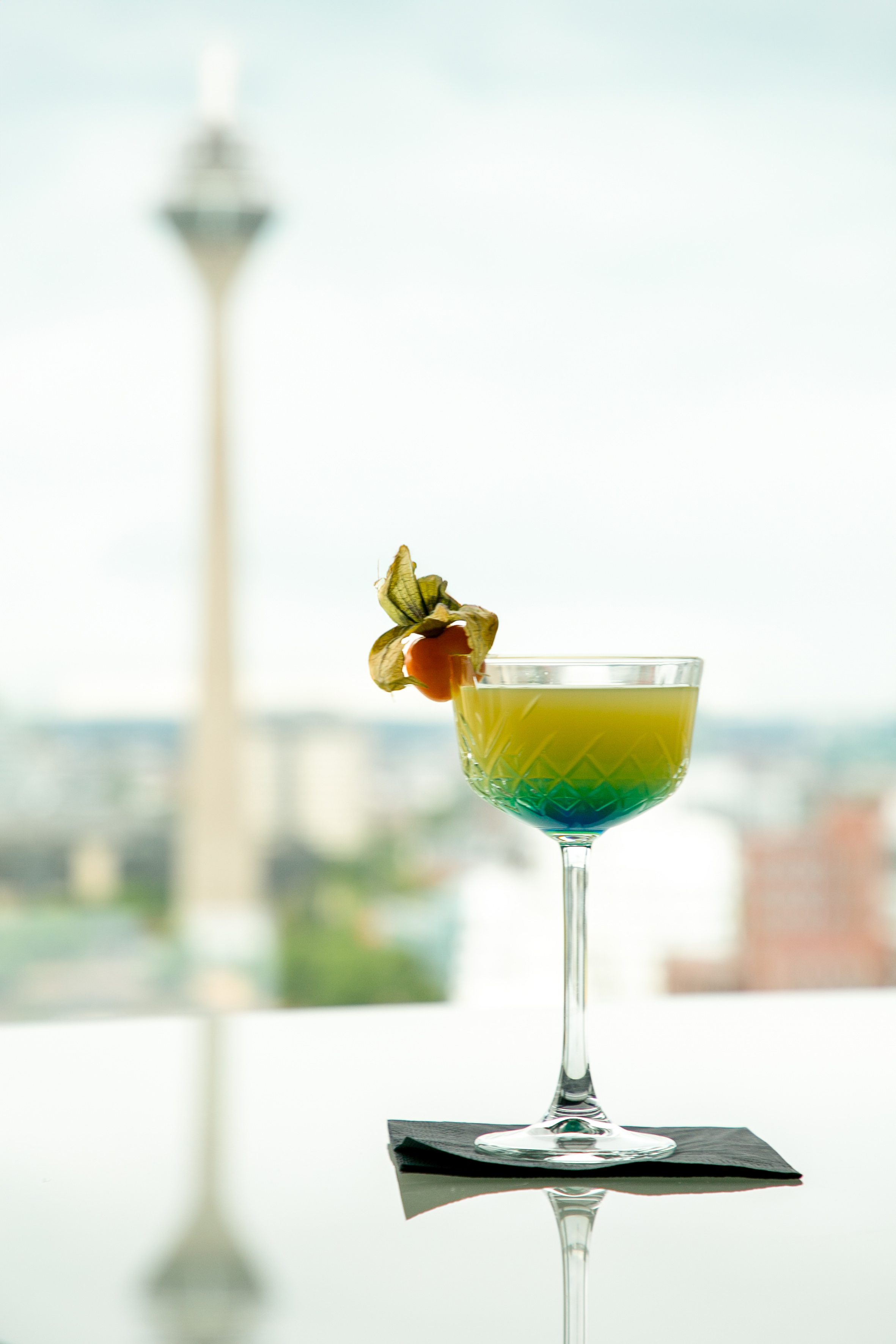 Gradient cocktail with physalis garnish, on table with blurred city tower view.