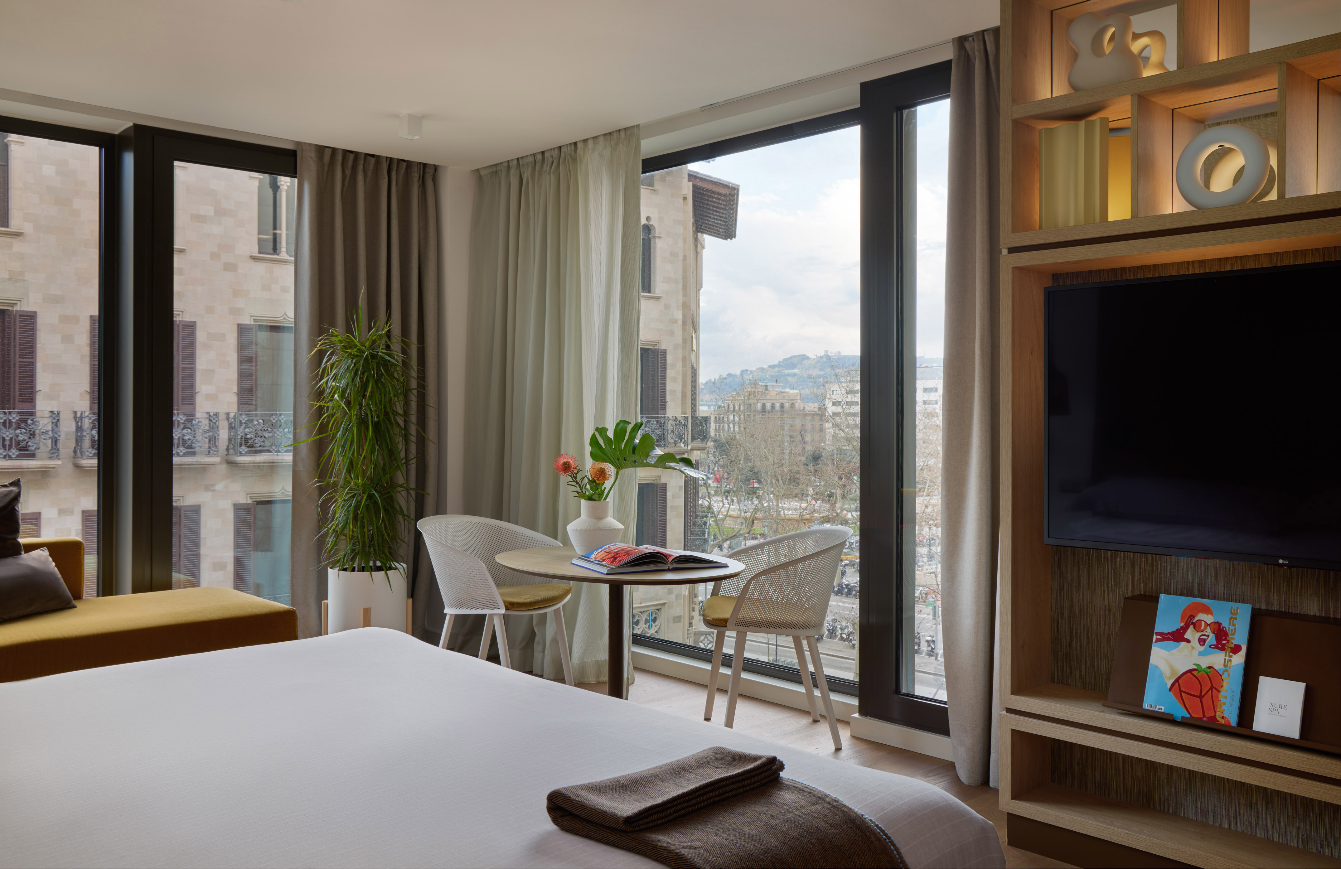 Modern hotel room with city view, large windows, comfortable bed, and dining table.
