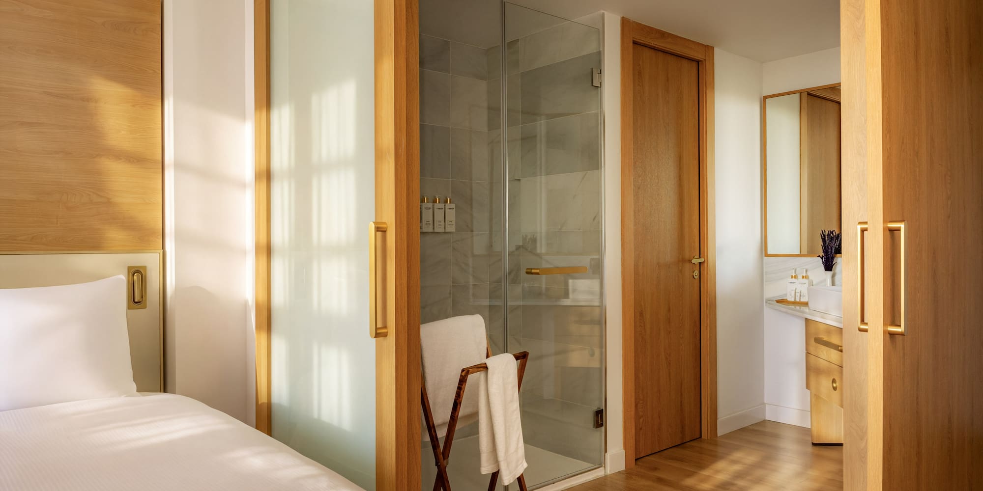 Modern hotel room with white bed, wood accents, glass shower, and bathroom vanity.