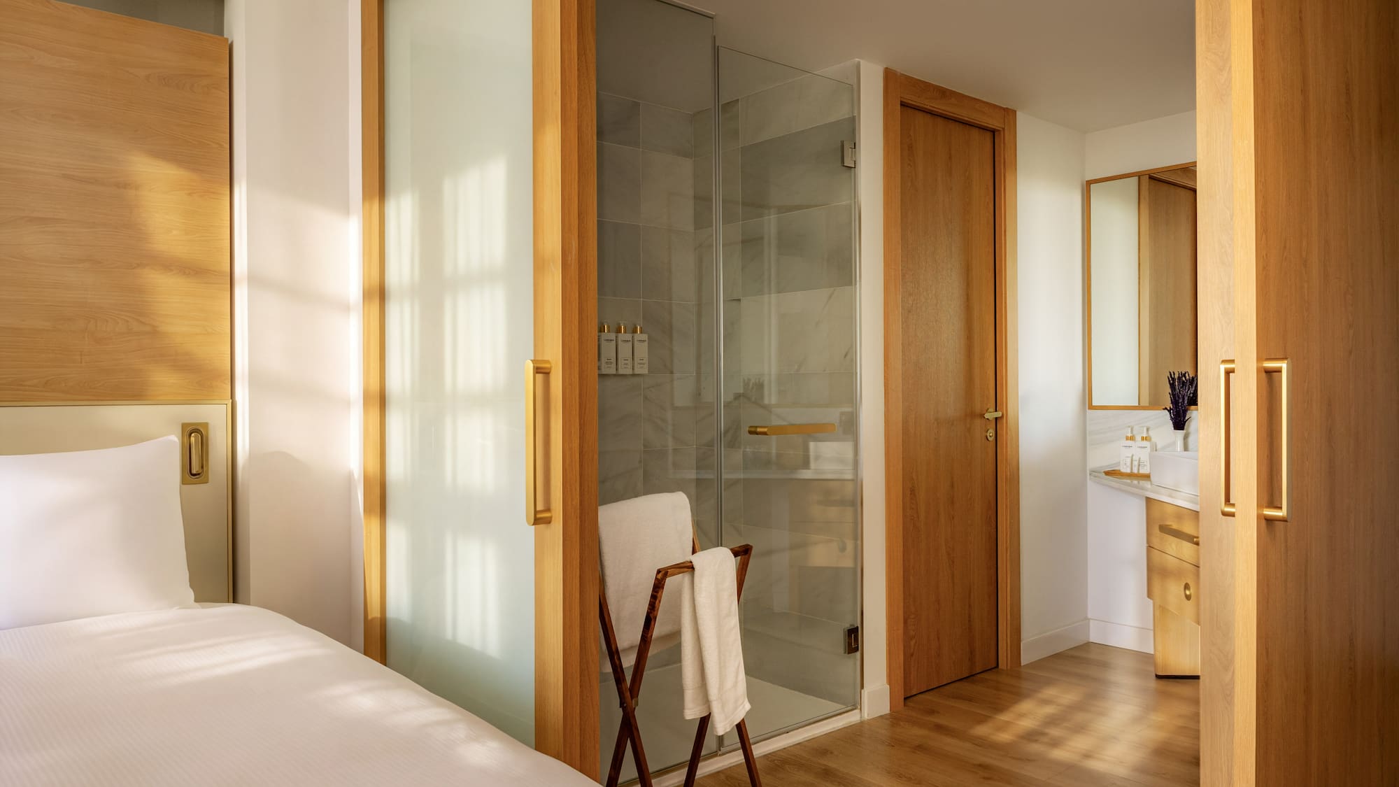 Modern hotel room with white bed, wood accents, glass shower, and bathroom vanity.