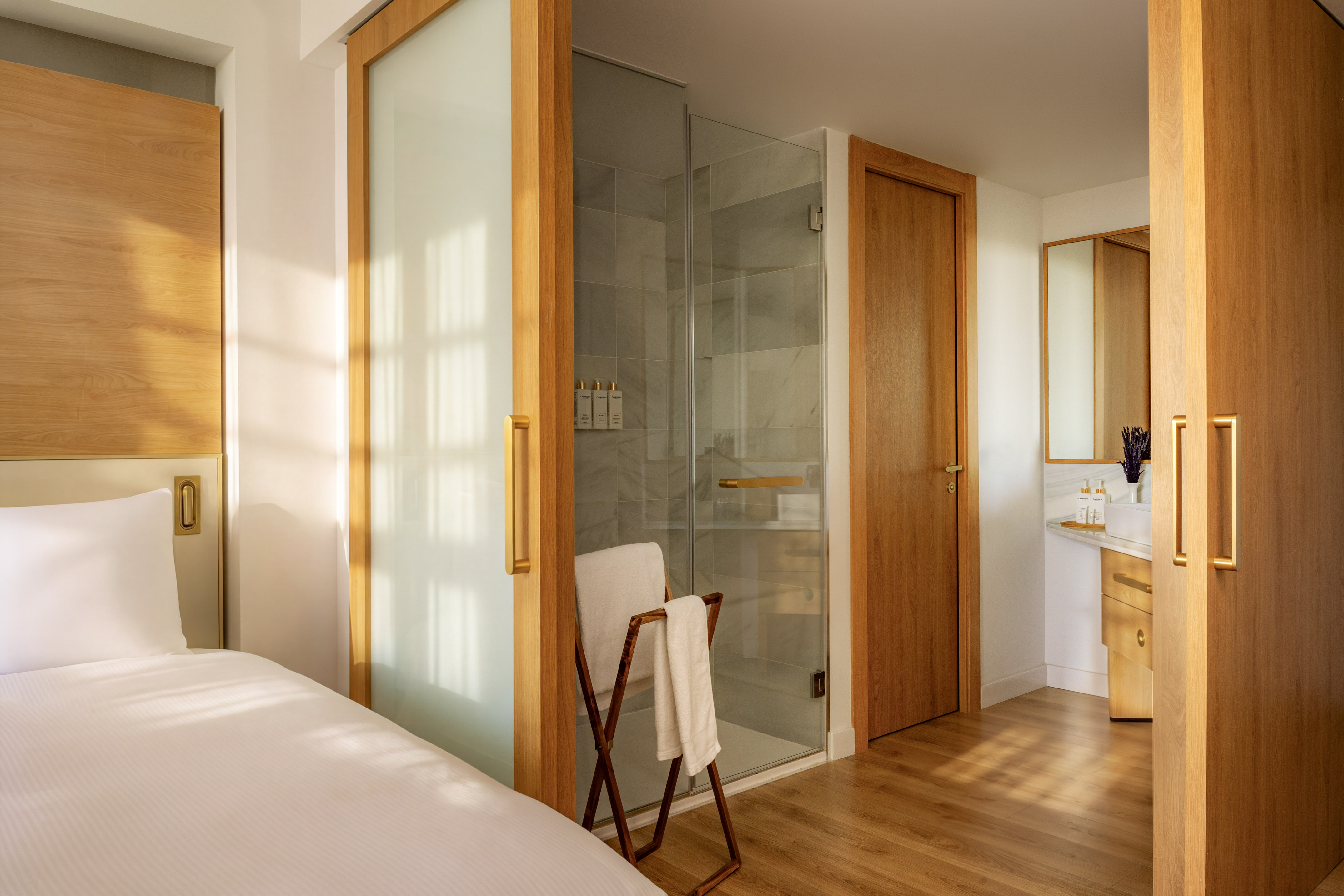 Modern hotel room with white bed, wood accents, glass shower, and bathroom vanity.