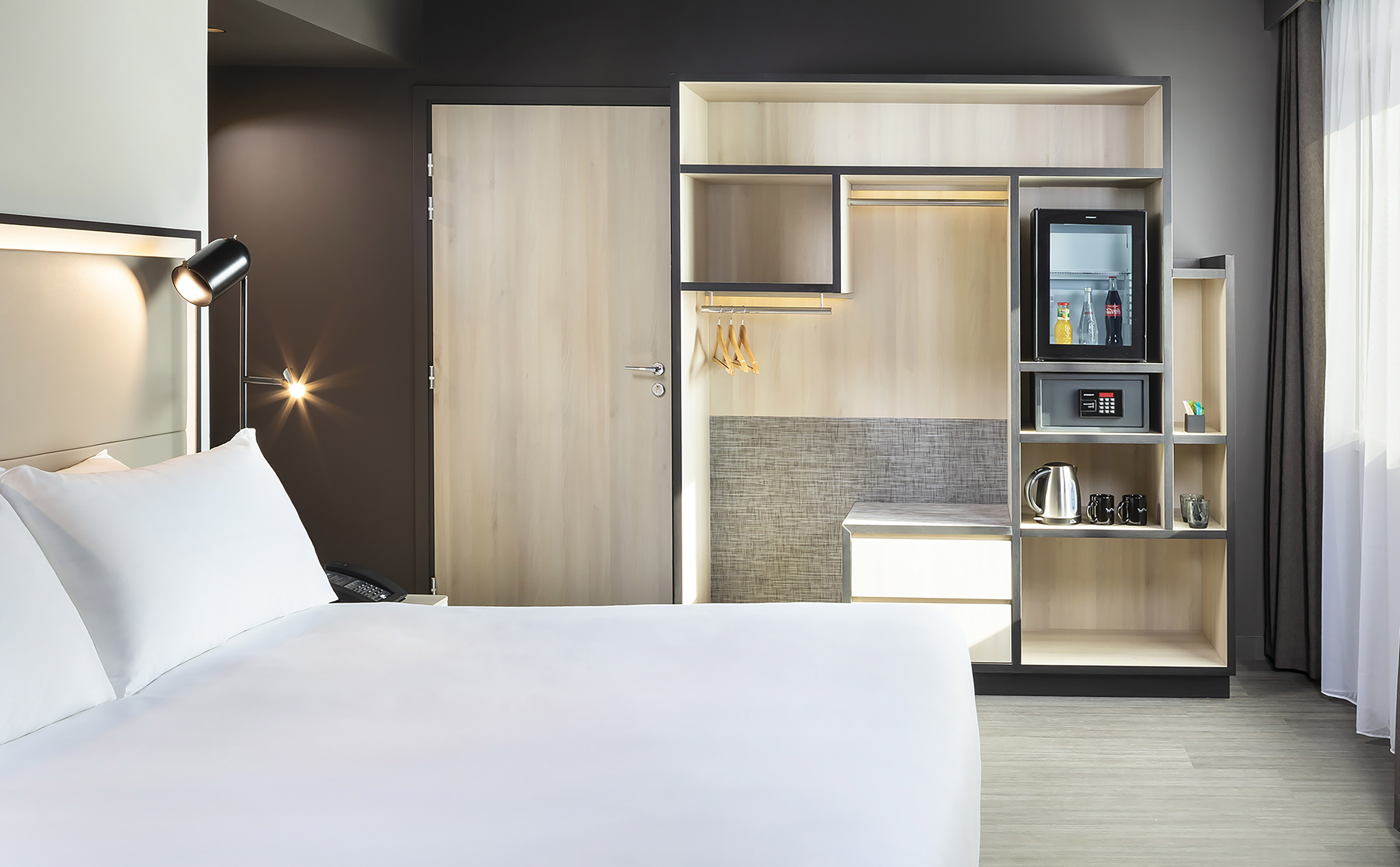 Modern hotel room with white bed, built-in closet, mini-fridge, and safe.