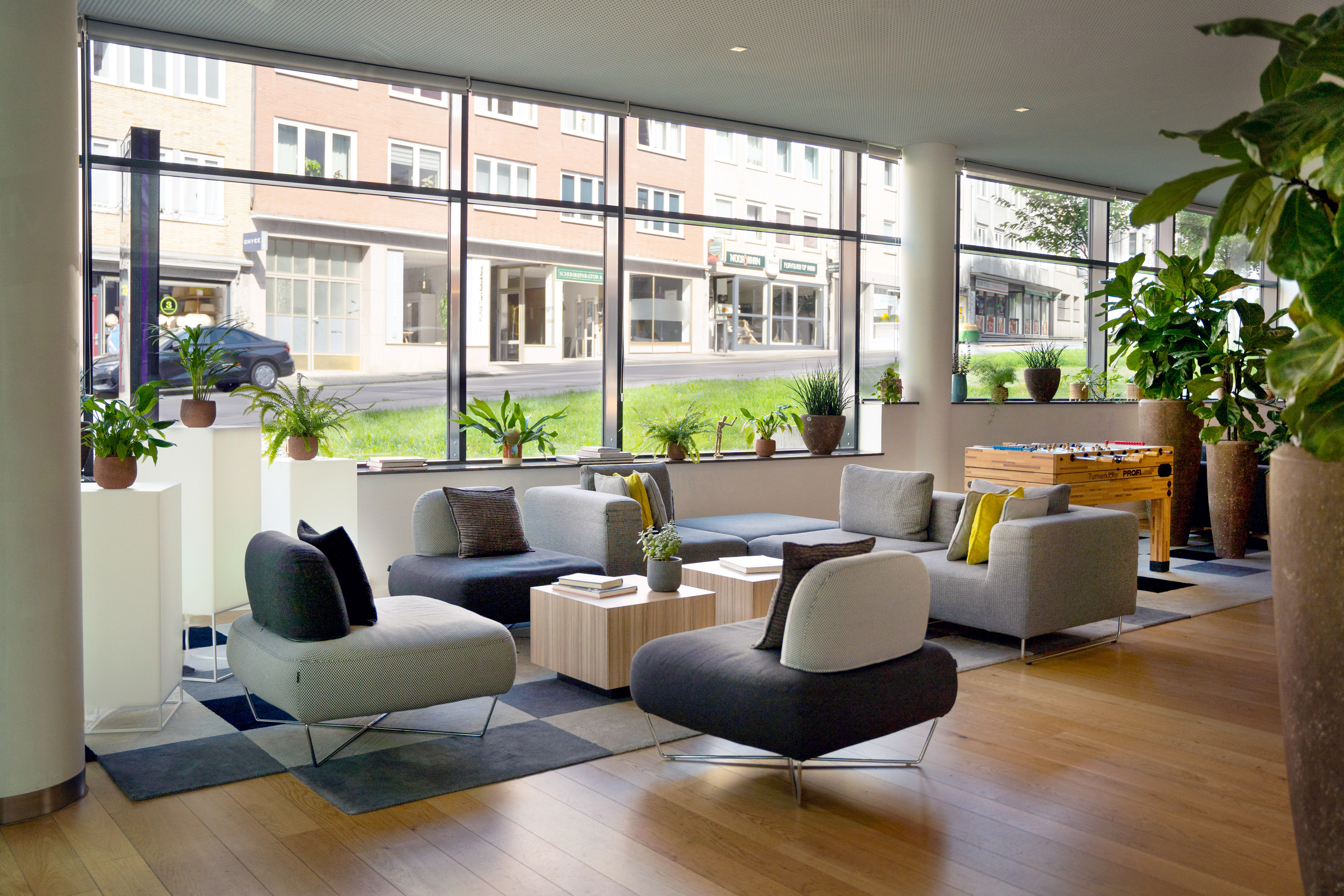 Modern lounge with grey seating, large windows, plants, and a foosball table.