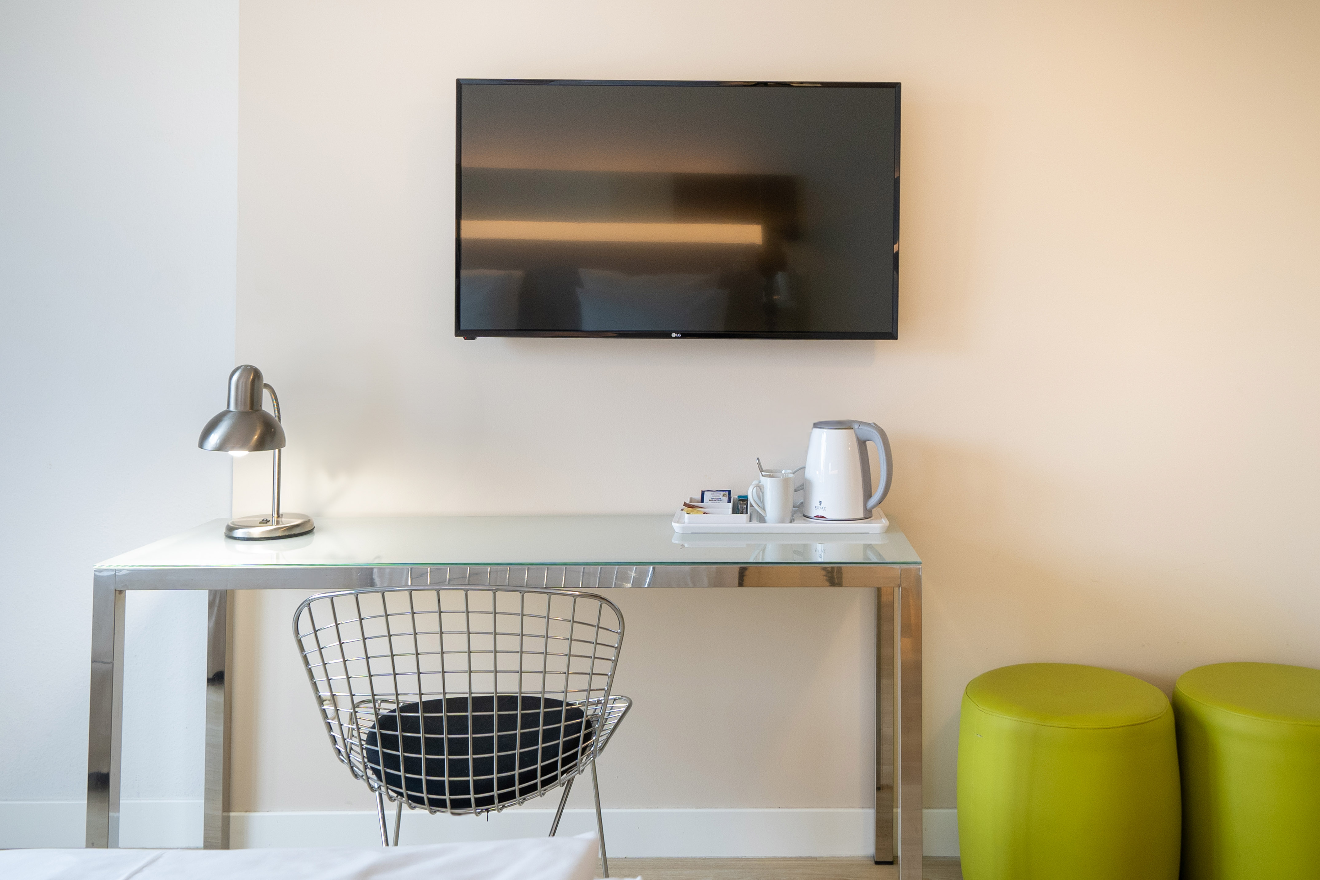 Modern hotel room with wall-mounted TV, glass desk, metal chair, and tea kettle.