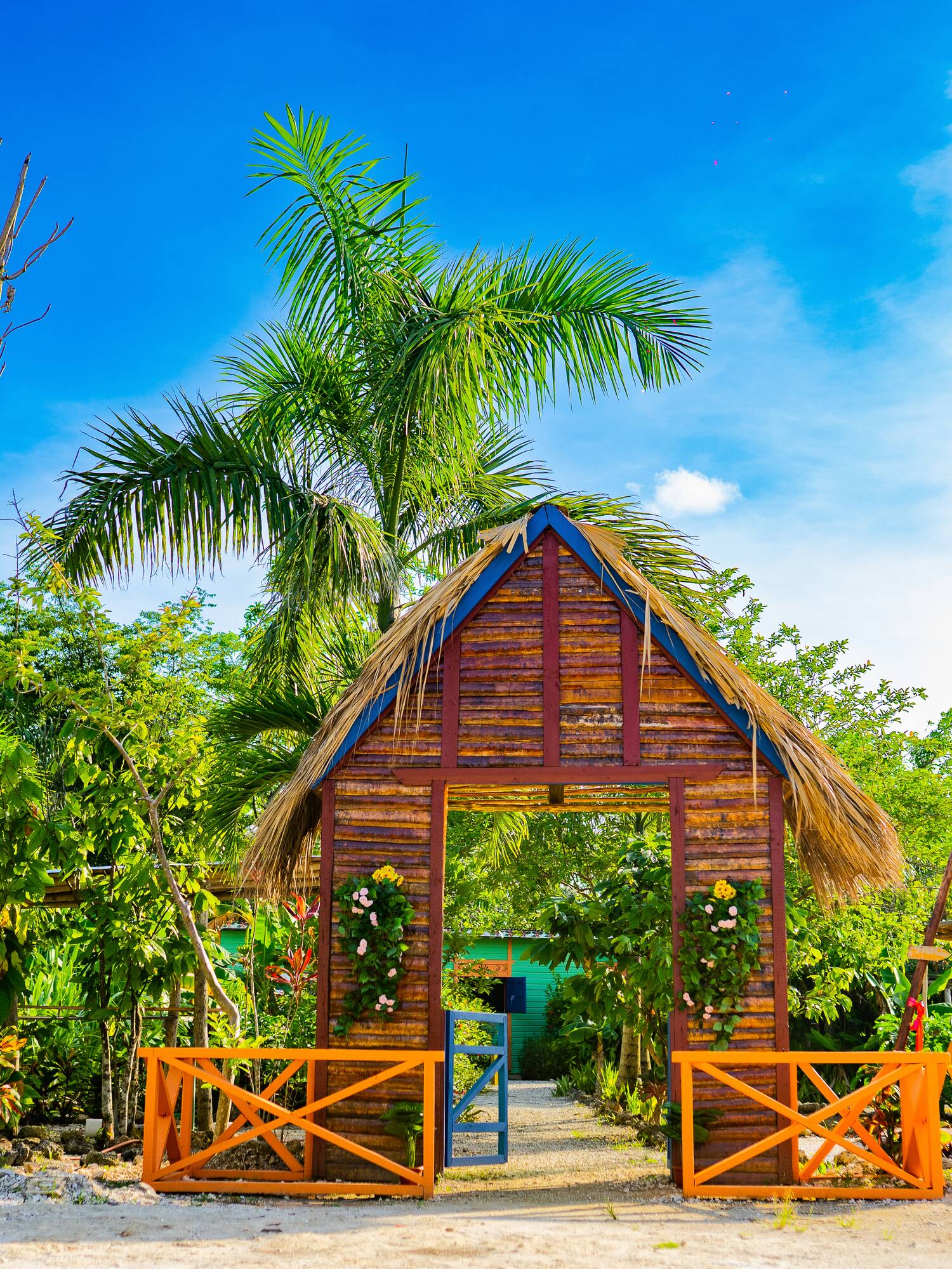 Wooden archway with thatched roof, orange gates, and tropical garden plants.