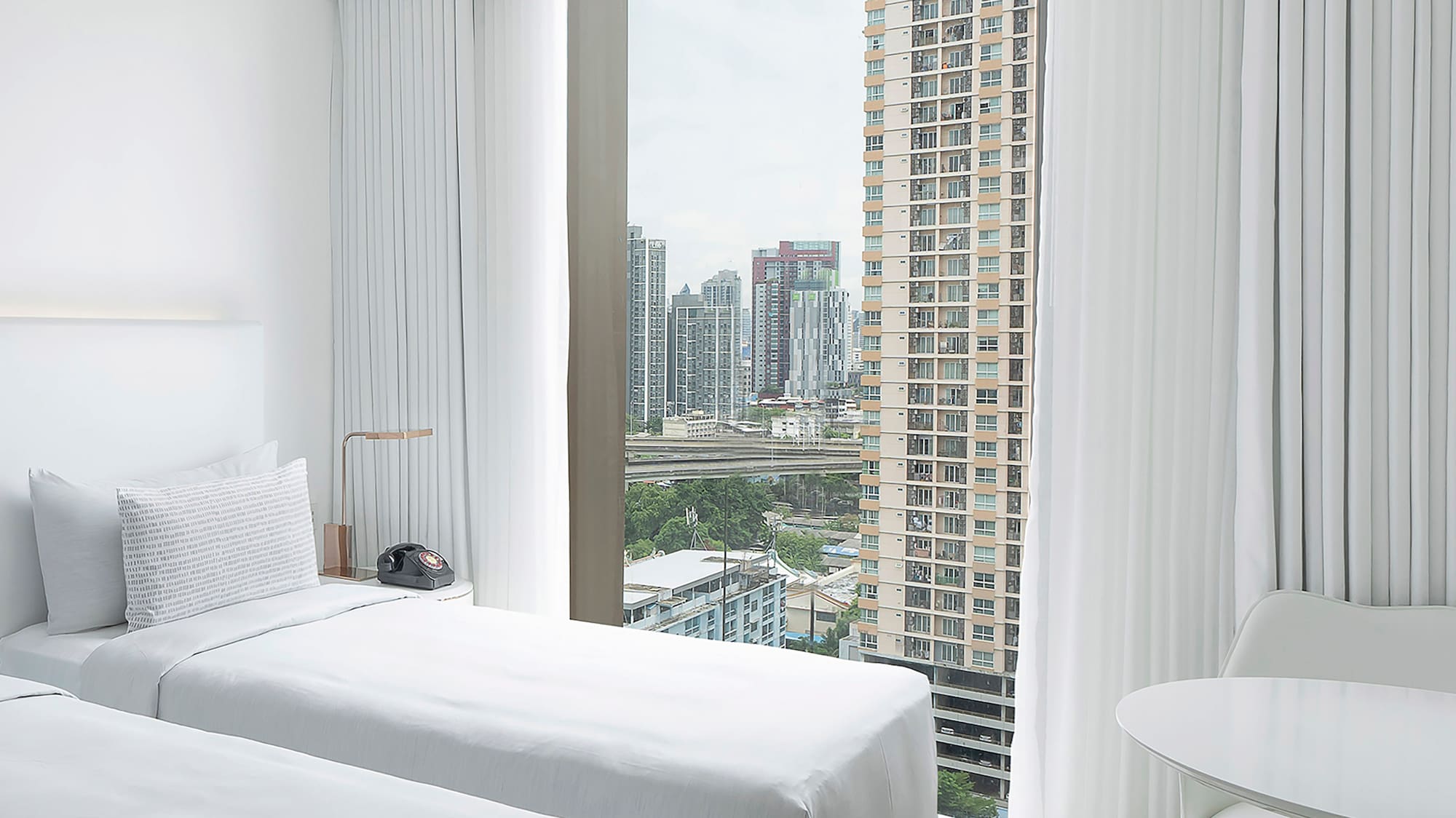 a room with two beds and a table and a window with tall buildings in the background