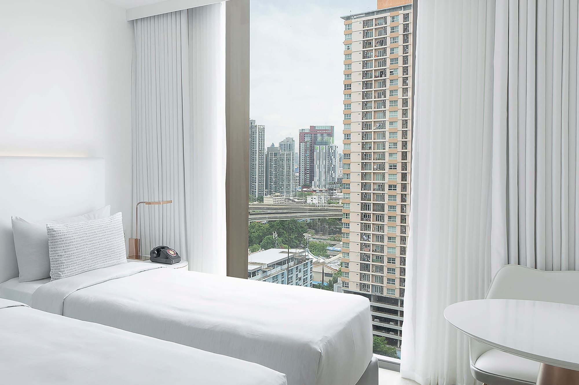a room with two beds and a table and a window with tall buildings in the background