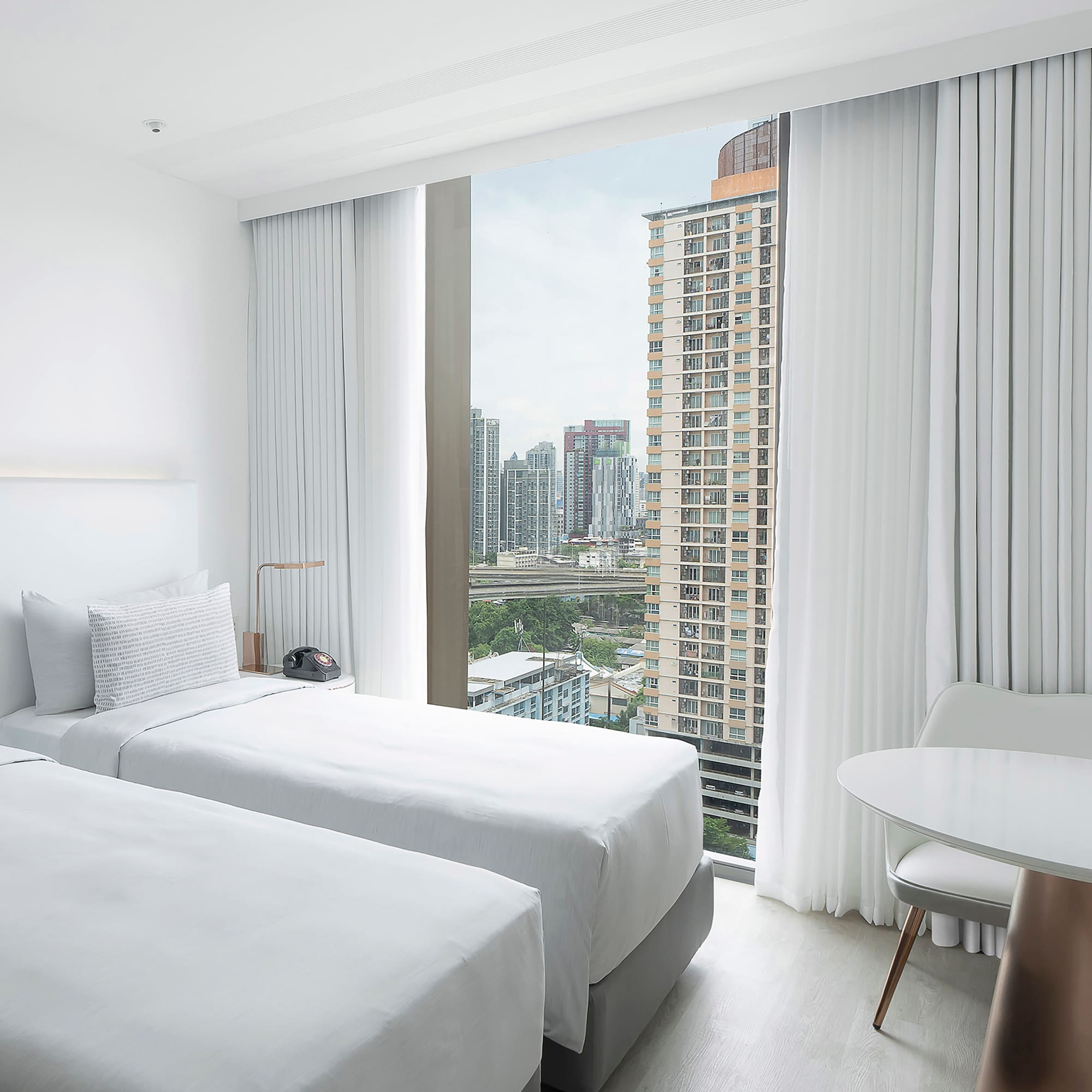 a room with two beds and a table and a window with tall buildings in the background