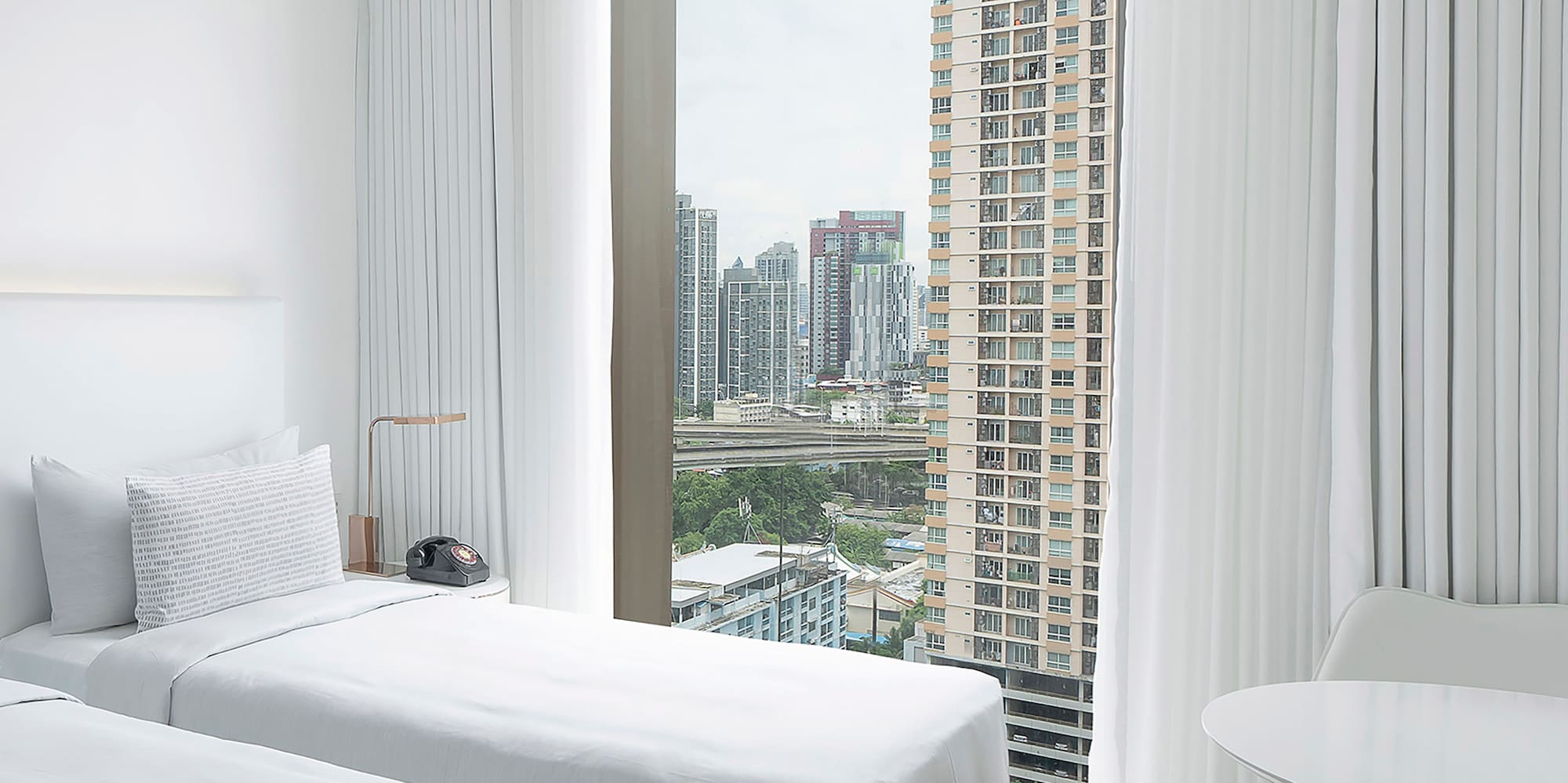 a room with two beds and a table and a window with tall buildings in the background